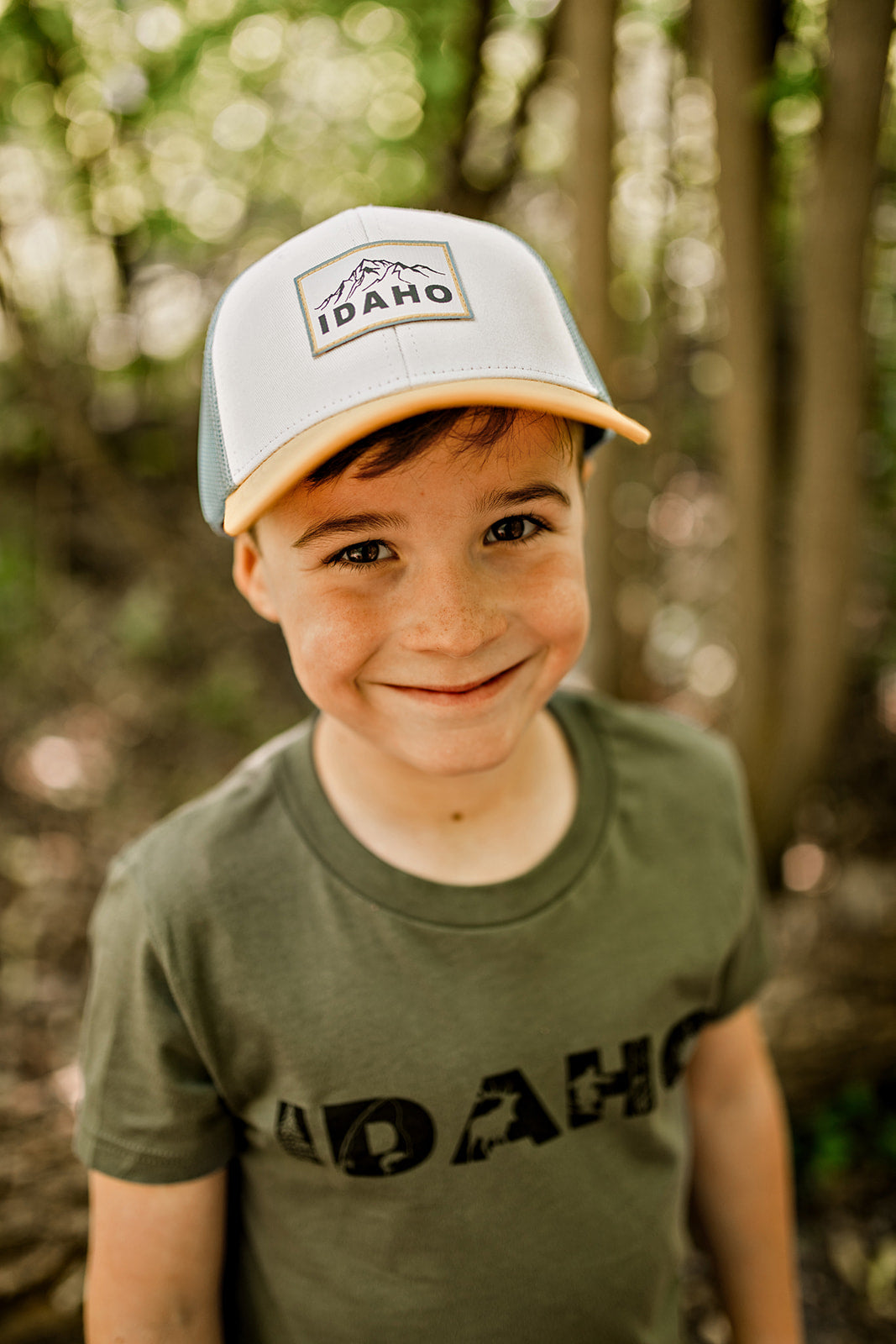 Smiling boy wearing an Idaho hat and T-shirt, standing in a sunlit forest area. His cheerful expression highlights the joy of exploring Idaho's natural beauty.