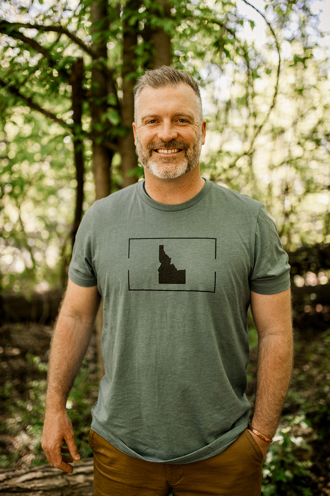 Smiling man outdoors wearing a green Idaho-themed t-shirt with a minimalist black Idaho state outline. Ideal for showcasing Idaho-inspired apparel for men, highlighting local pride and style in nature setting