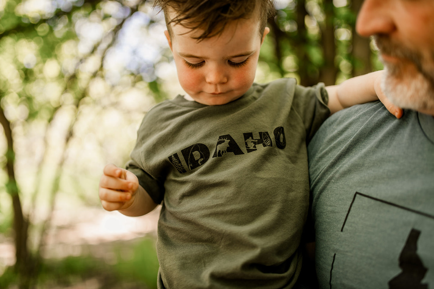 The Idaho T shirt comes in military green, and is being worn by a toddler. He is wearing a T shirt that says Idaho on it. Inside of the letters are different graphics that represent Idaho.