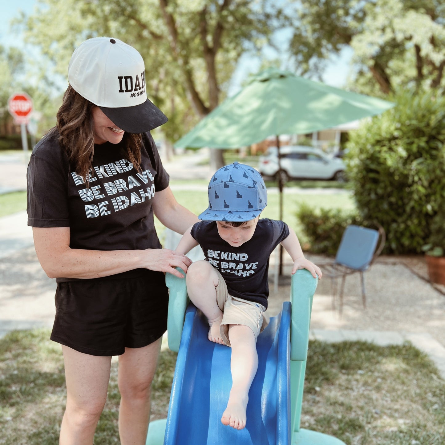 Mom and child wearing matching "Be Kind, Be Brave, Be Idaho" tees and TatorJo hats; mom wears a black and cream Idaho Mama Hat and child wears a blue Idaho Hat with mountain design captured outdoors on a sunny Idaho day. Idaho Hats designed by TatorJo