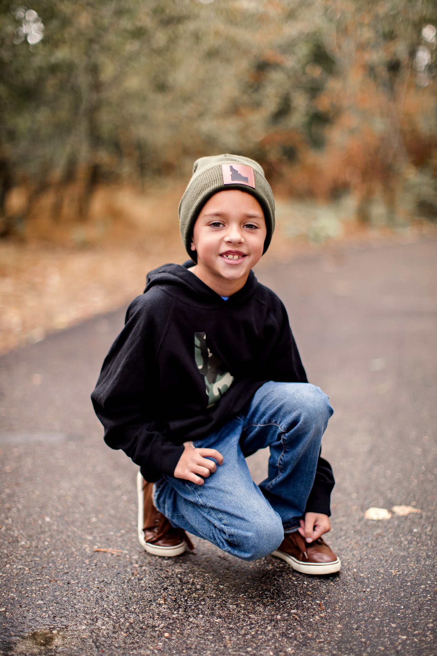 This black camo hoodie has an Idaho printed on it. A young boy is wearing an Idaho hat, beanie, and Idaho clothing showing off his Idaho roots. The boy is outside in Idaho enjoying the great outdoors.