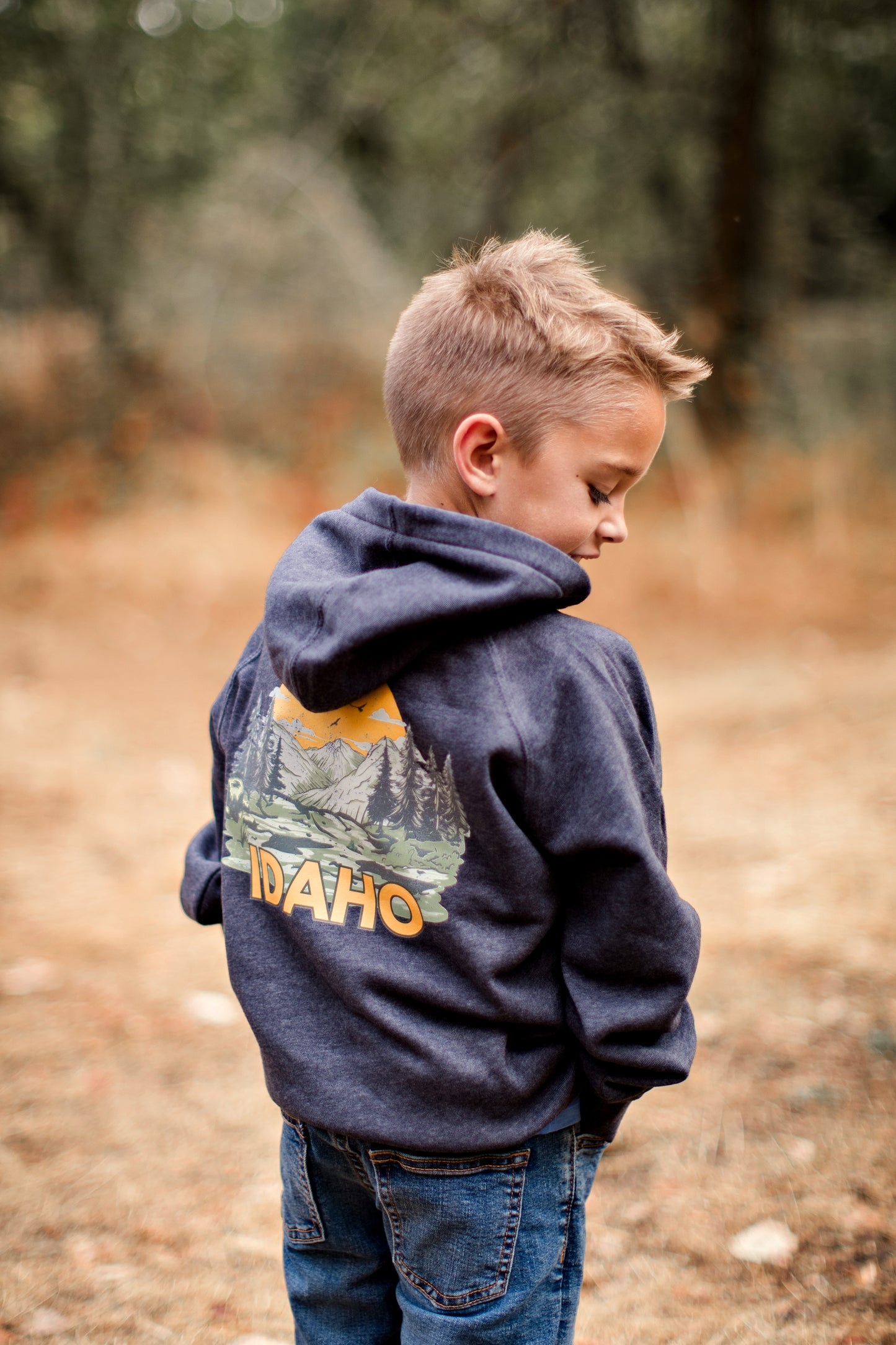 A young boy outside in Idaho wearing an Idaho sweater. This sweater is blue with a big Idaho design on the back and front corner pocket. The Idaho design has Idaho mountains on it and the wording Idaho. This sweater is made for toddlers, youth, and adults. This sweater is designed by TatorJo, an Idaho Clothing Company