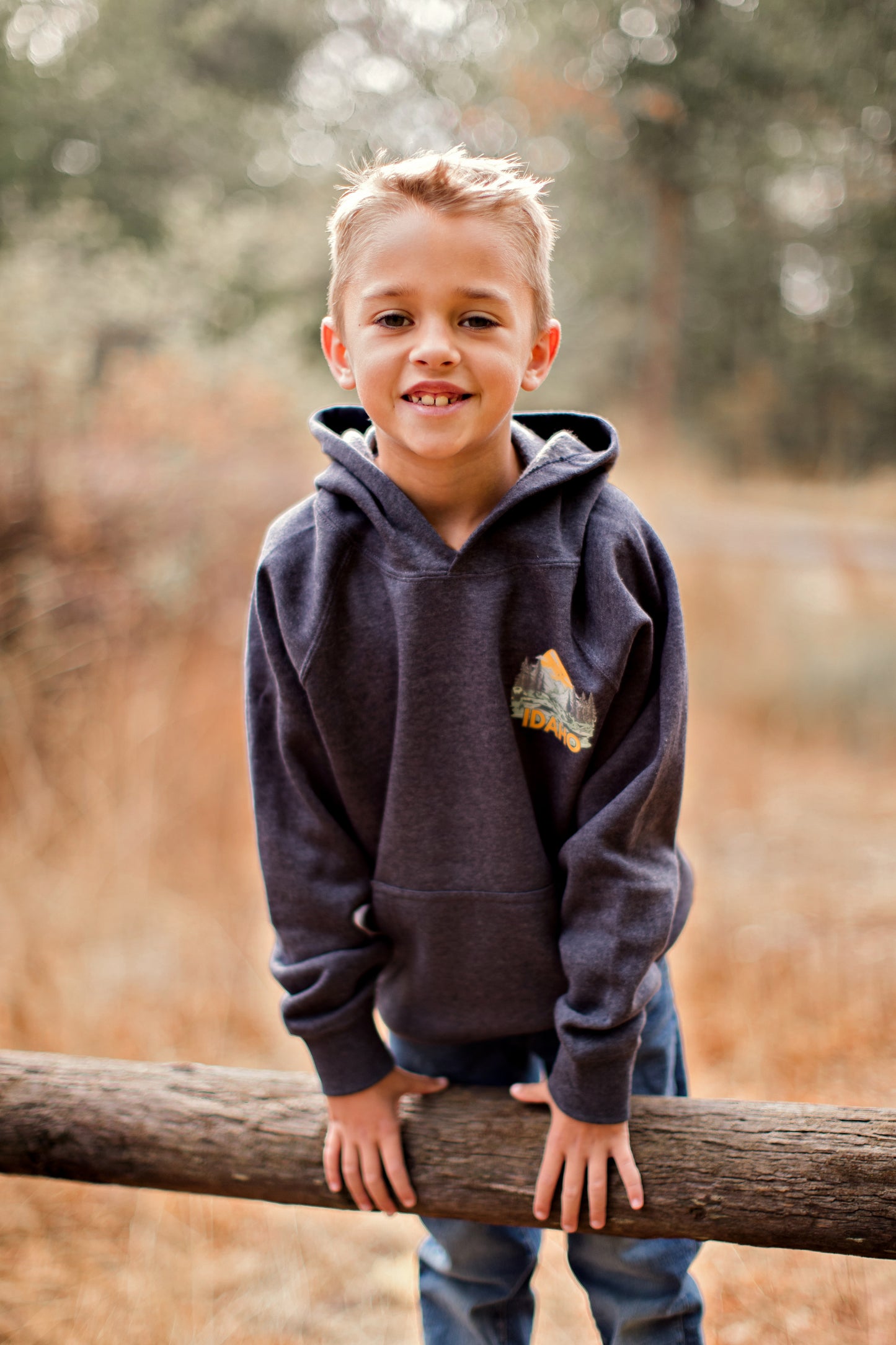 A young boy outside in Idaho wearing an Idaho sweater. This sweater is blue with a big Idaho design on the back and front corner pocket. The Idaho design has Idaho mountains on it and the wording Idaho. This sweater is made for toddlers, youth, and adults. This sweater is designed by TatorJo, an Idaho Clothing Company