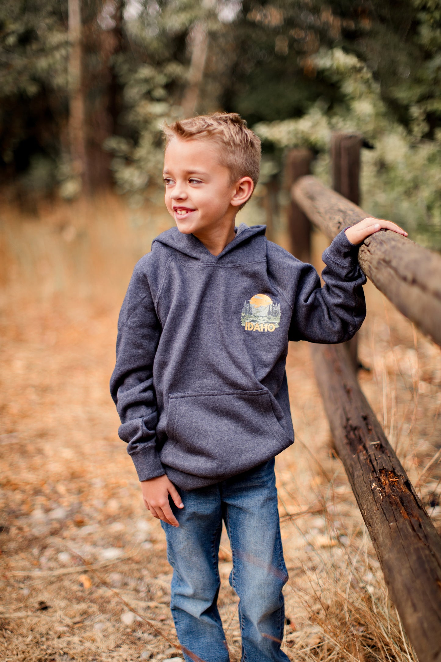 A young Idaho boy outside standing in his Idaho sweatshirt. The sweatshirt is blue and has a big Idaho design on it. The Idaho design has Idaho mountains on it. This Idaho sweater is designed by TatorJo, an Idaho Clothing Company.