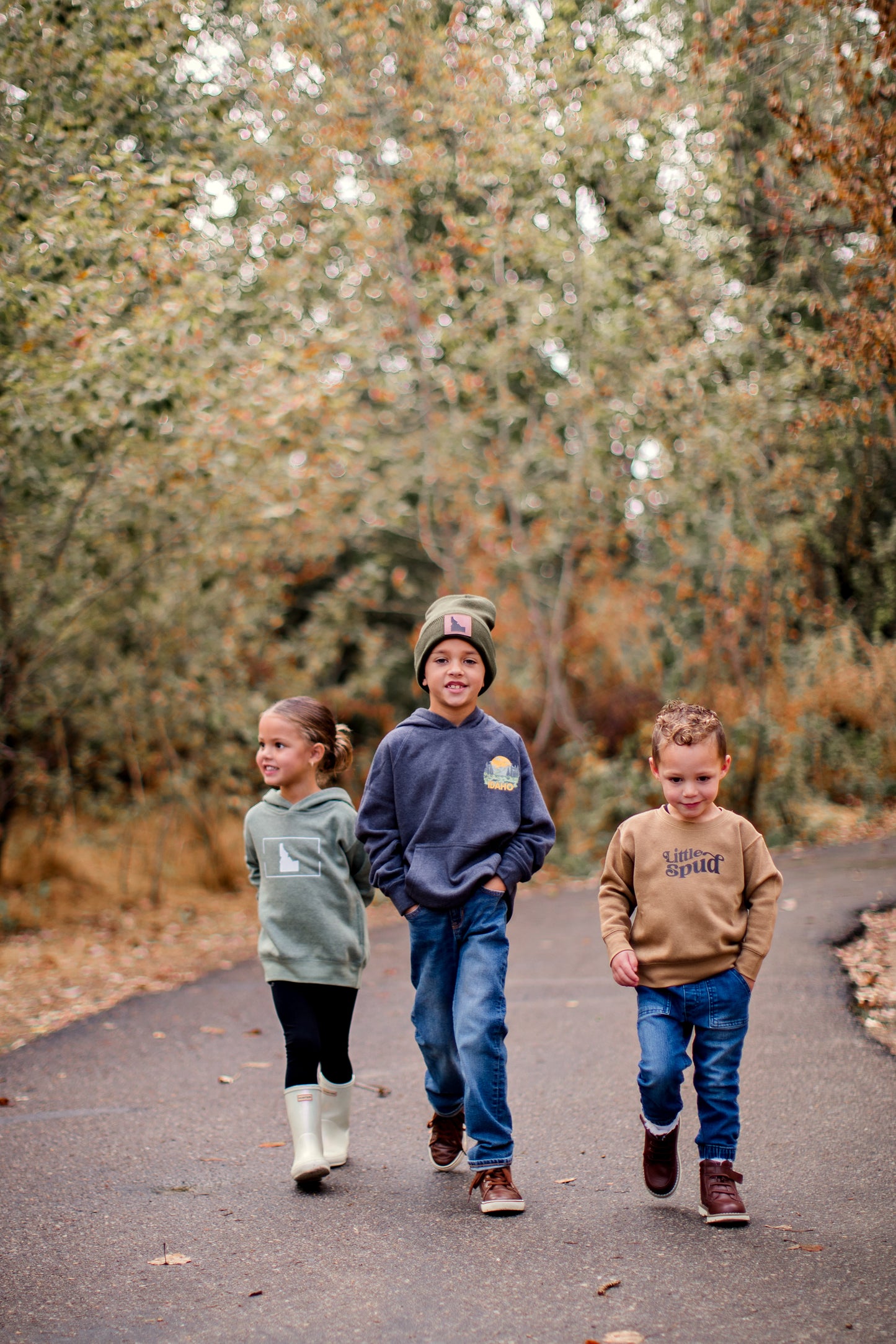Idaho kids standing outside in their TatorJo apparel. Each kid has an Idaho sweatshirt. The little girl on the left has an Idaho sweater that is green. The middle boy has a blue sweater with an Idaho mountain design on it. The little boy on the right has a brown sweater on that says Little Spud. All kids are wearing Idaho Clothing.