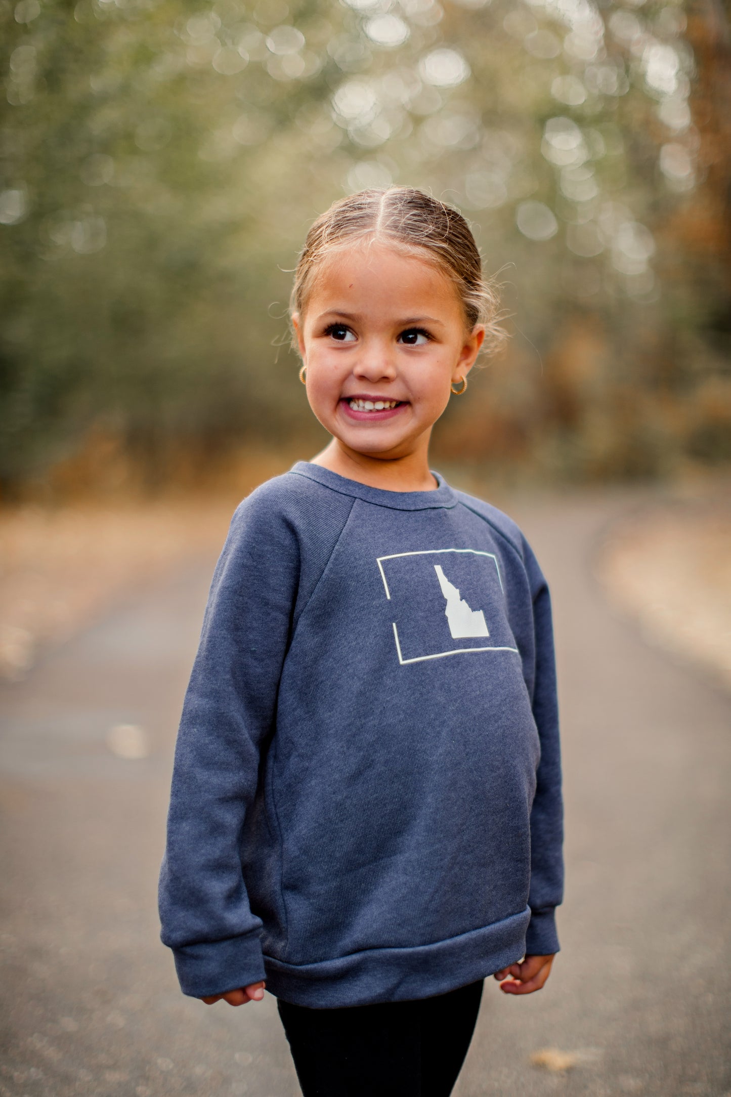 A little girl wearing a blue crewneck with a simple Idaho design on it. The little girl is standing outside in Idaho.