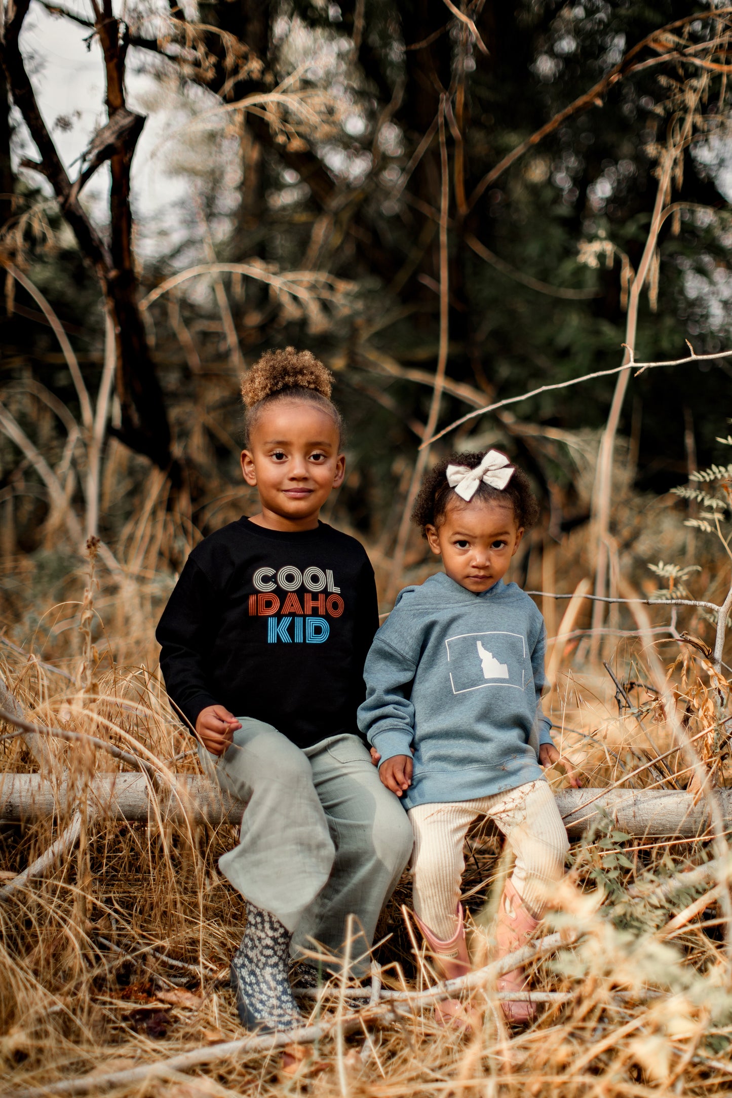 Two little girls sitting outside in their Idaho clothing. One little girl is wearing an Idaho shirt that says Cool Idaho Kid. The other little girl is wearing a blue sweatshirt with an Idaho design on it. These clothes are designed in Idaho by TatorJo, an Idaho Clothing Company.
