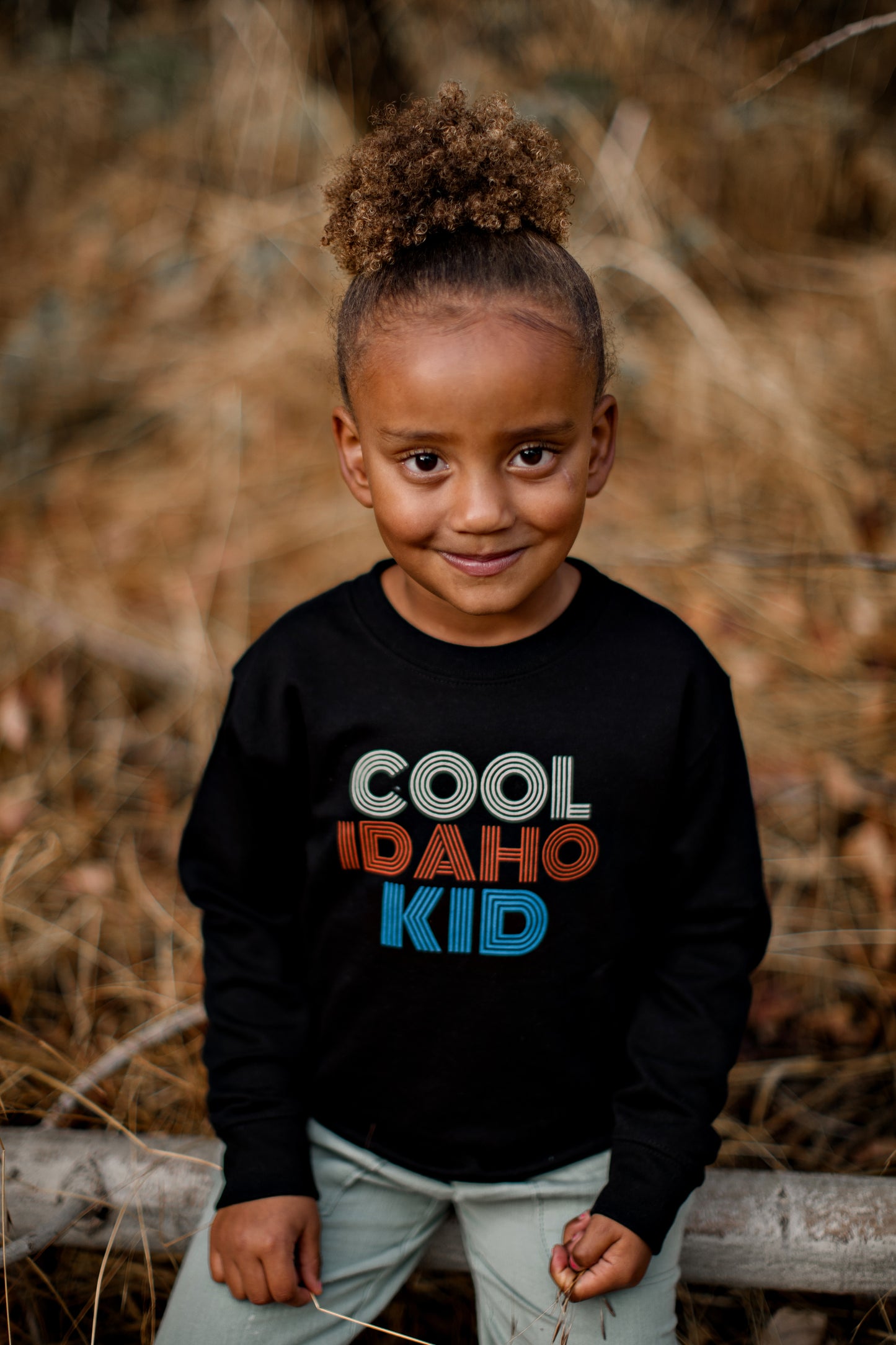 A little girl outside wearing an Idaho sweater. The Idaho sweater is black and says Cool Idaho Kid written on it. The sweater is designed by TatorJo an Idaho Apparel company in Idaho.