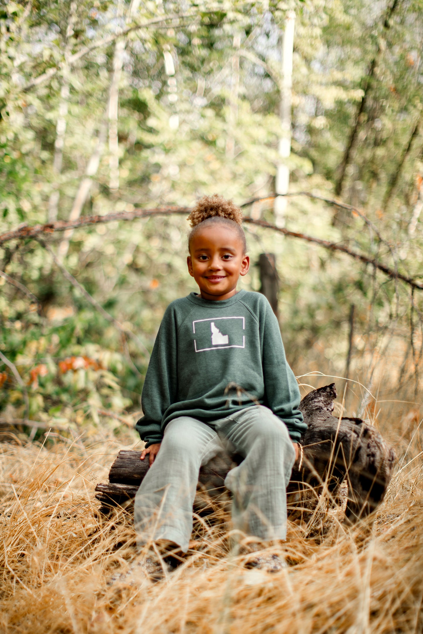 A little girl sitting outside in Idaho wearing an Idaho sweater. The Idaho sweater has a simple Idaho design on it in white. This shirt shows off Idaho pride.