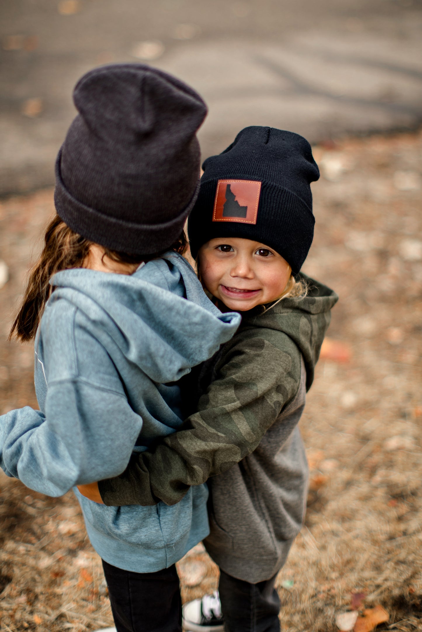 Two siblings standing outside in the fall. They are both wearing Idaho beanies. One is wearing a Black Idaho Hat, that is a warm beanie. The little Idaho girl is wearing a gray idaho beanie. Each one represents Idaho. Idaho living!