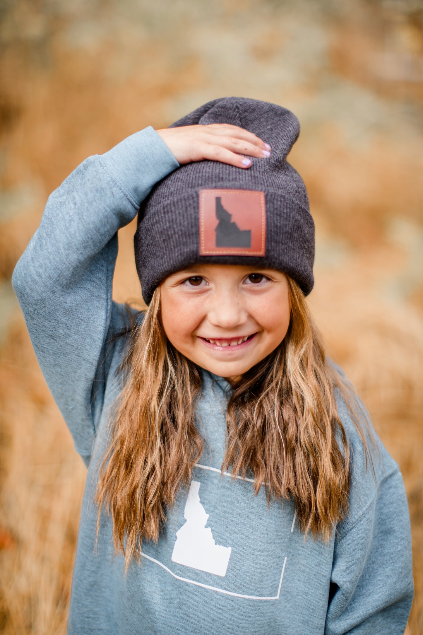 A young girl standing outside in Idaho wearing an Idaho beanie. The Idaho beanie has a leather patch on it. The patch has Idaho on it. The Idaho beanie is gray