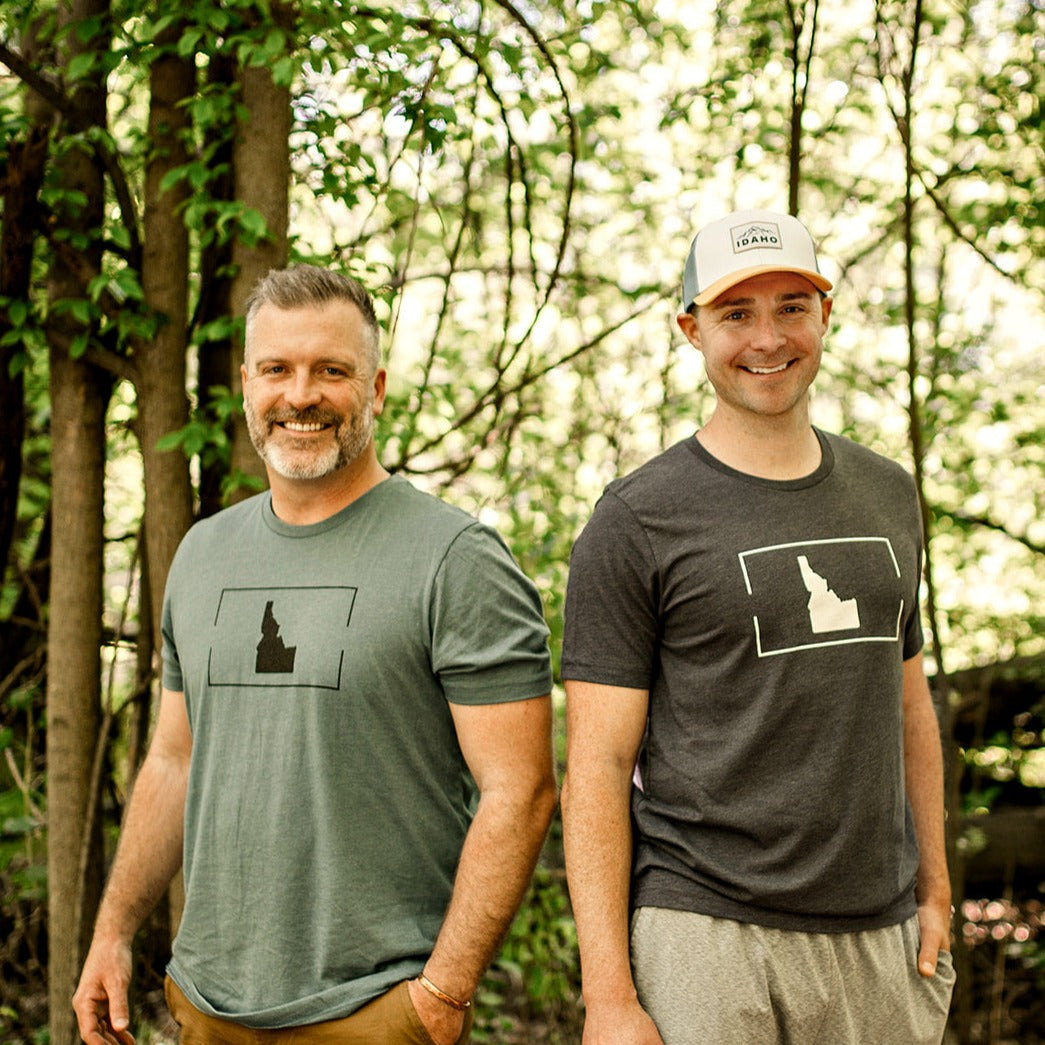 Two men standing outside in Idaho. The men are wearing a blue Idaho shirt and a gray Idaho shirt. The shirts have a simple Idaho design on them. The Idaho T shirts are simple and clean looking. Showing off their Idaho pride