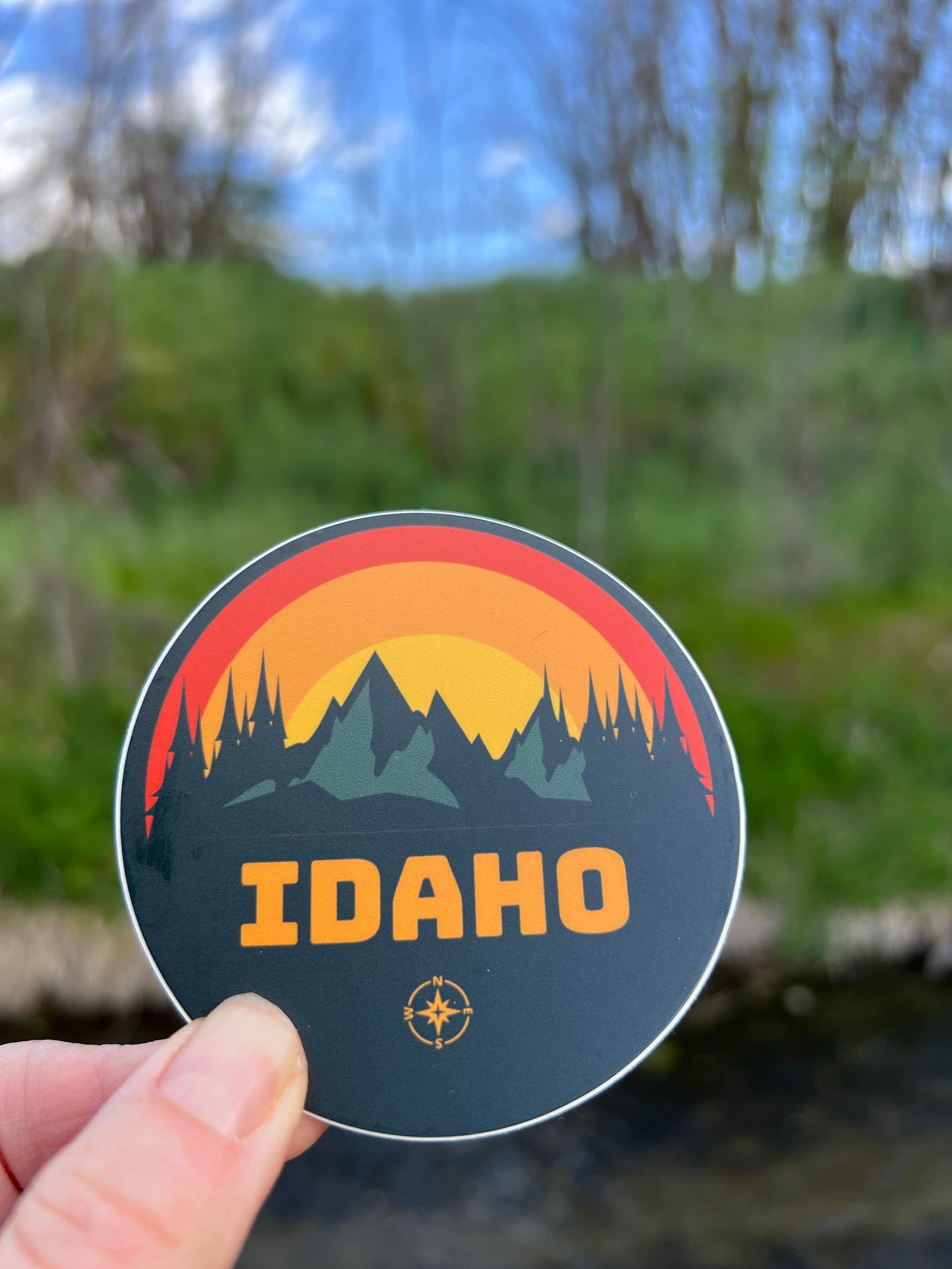 This Idaho Sticker has rugged mountains on it with a sunrise in the background. The sticker has Idaho written on it.