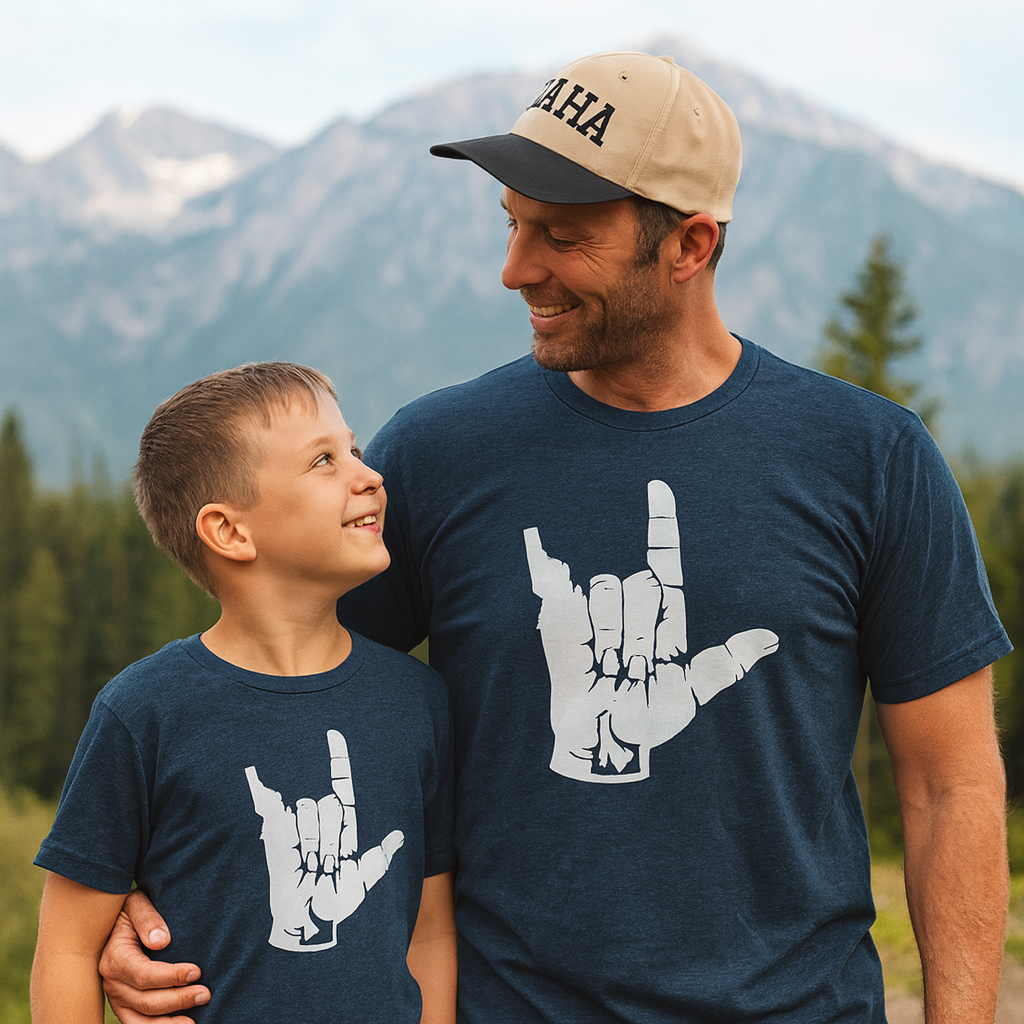 Man and child wearing matching navy t-shirts with hand sign graphics, standing in front of a mountainous landscape.