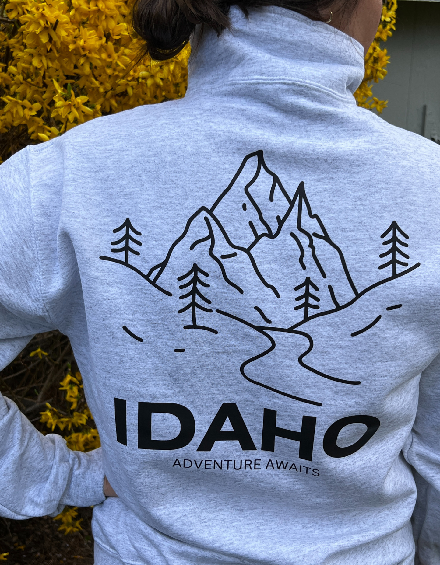 A person wearing a gray quarter zip sweater with 'IDAHO ADVENTURE AWAITS' printed on the back.