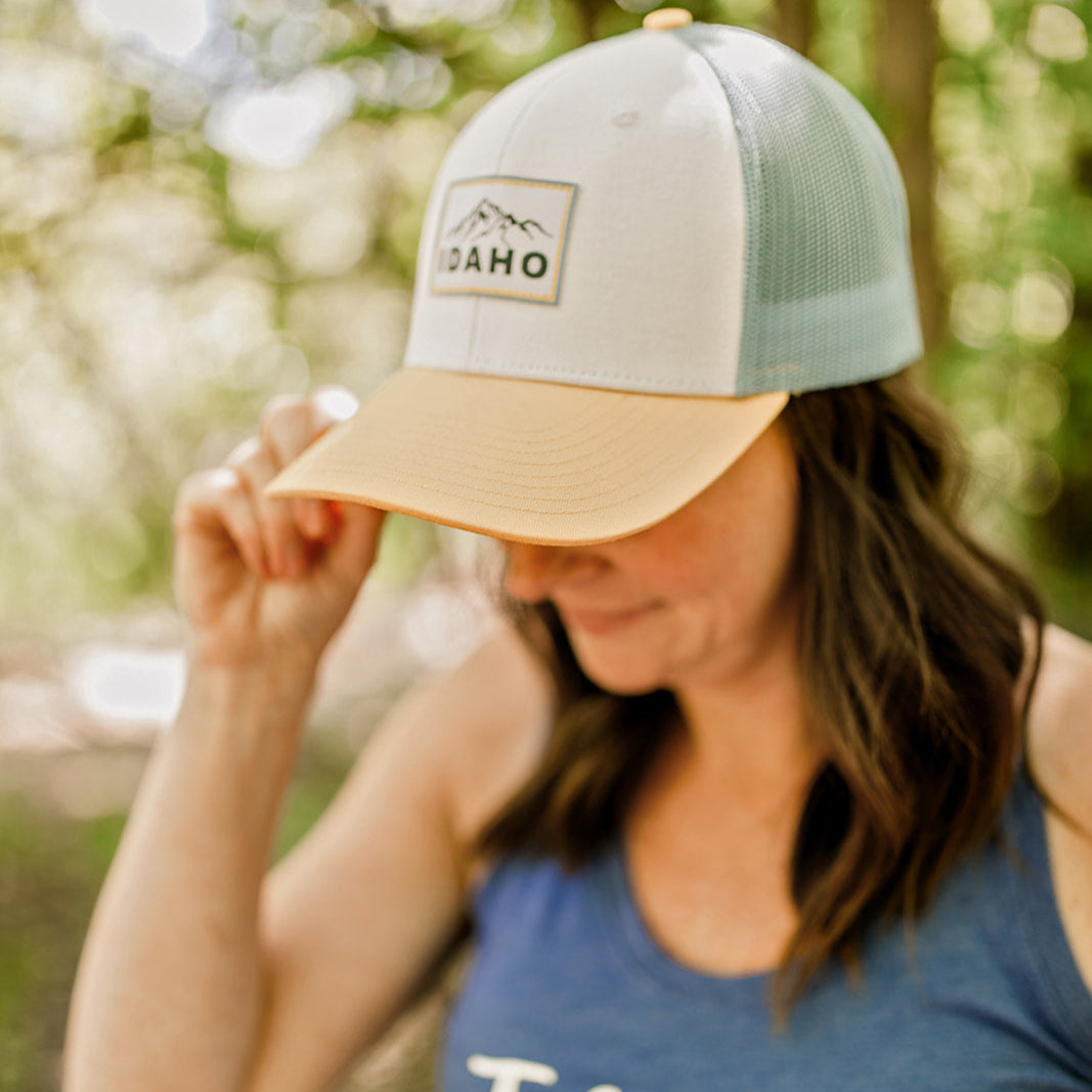 An Idaho mom wearing an Idaho Hat by TatorJo. The hat is yellow and blue and has the word Idaho on it. It has an Idaho mountain on it, and is a trucker style hat. This hat is designed by TatorJo, an Idaho apaprel company