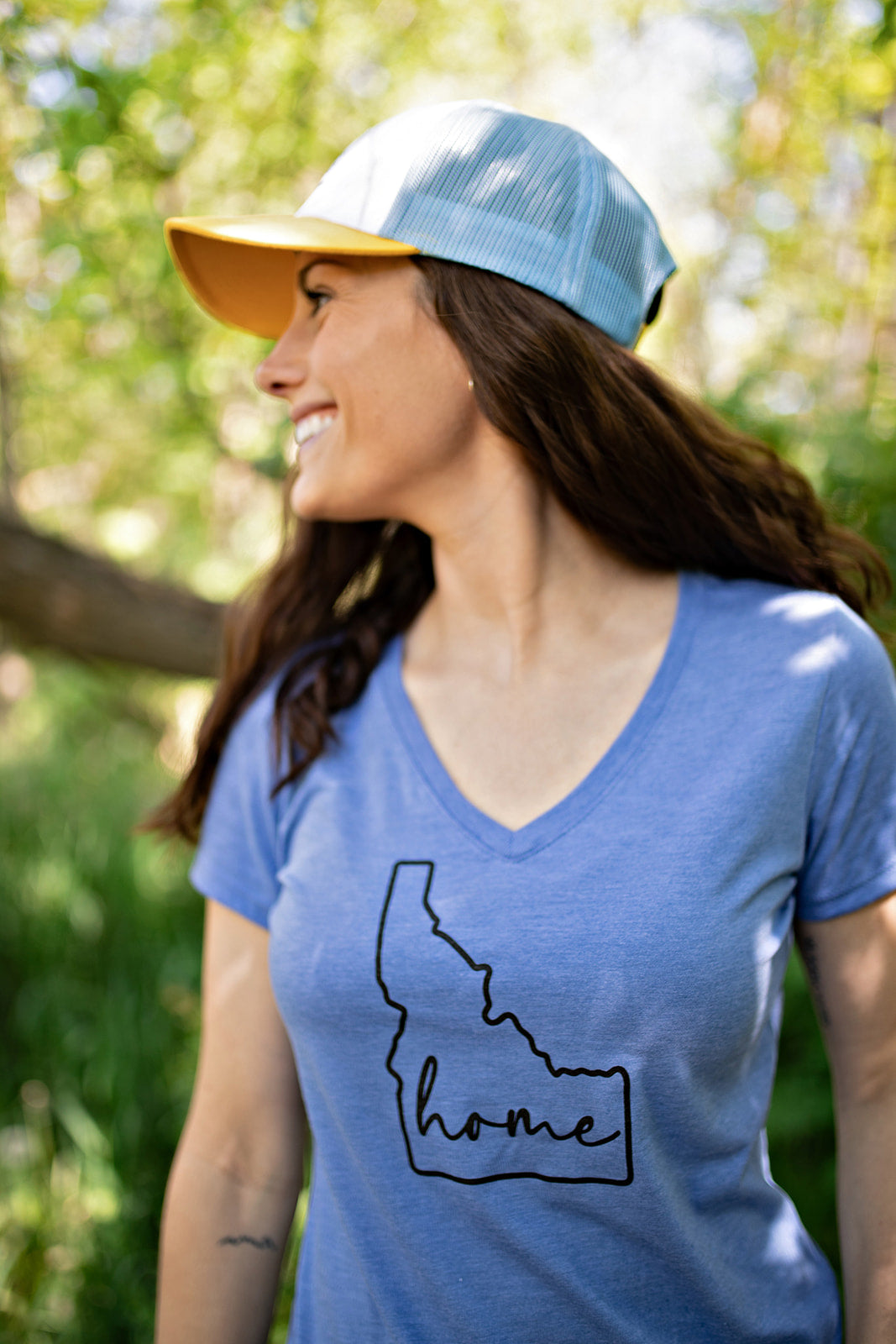 A young mom wearing an Idaho Trucker Hat. This Idaho Hat has Idaho mountains on it and the tag that says TatorJo. This hat comes in yellow and teal, with a bold mountain design on the front. Designed by TatorJo