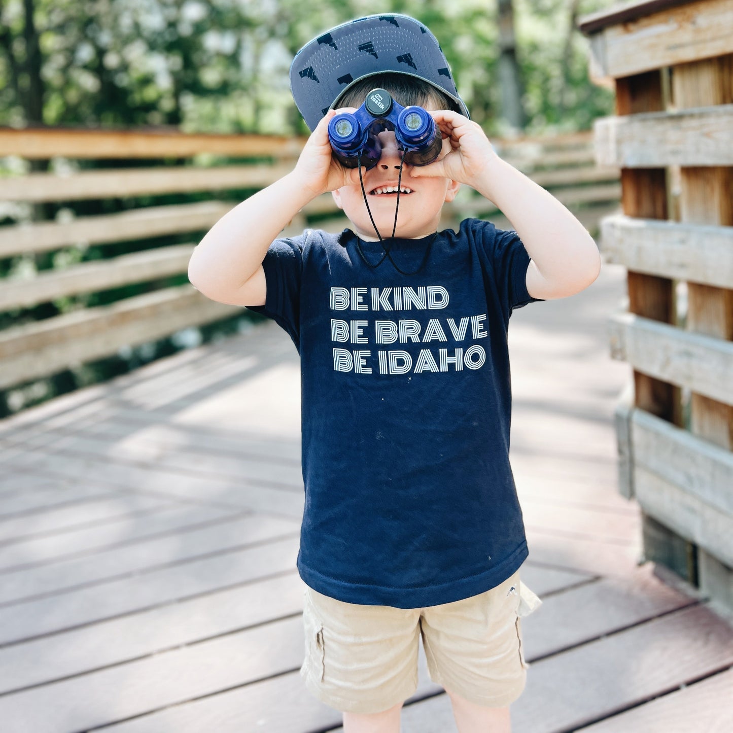 A little boy wearing an Idaho TatorJo T Shirt that says Be Kind, Be Brave, Be Idaho on it. The little boy is wearing an Idaho TatorJo hat. This shirt is designed by TatorJo, an Idaho apparel company.