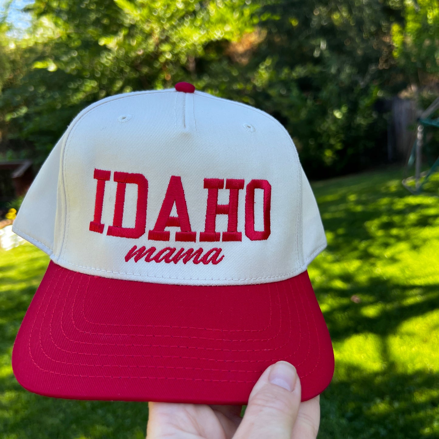 Red and white cap with 'Idaho Mama' text held by a hand against a blurred green outdoor background