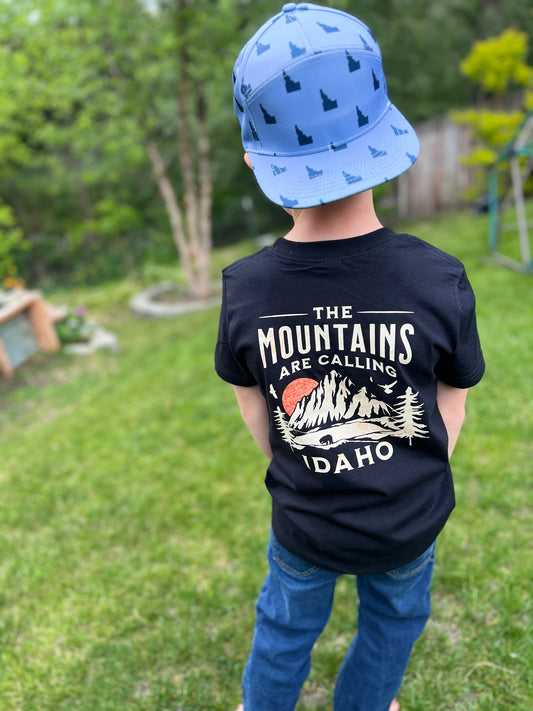 Child wearing a black t-shirt with 'The Mountains Are Calling Idaho' design, standing outdoors.