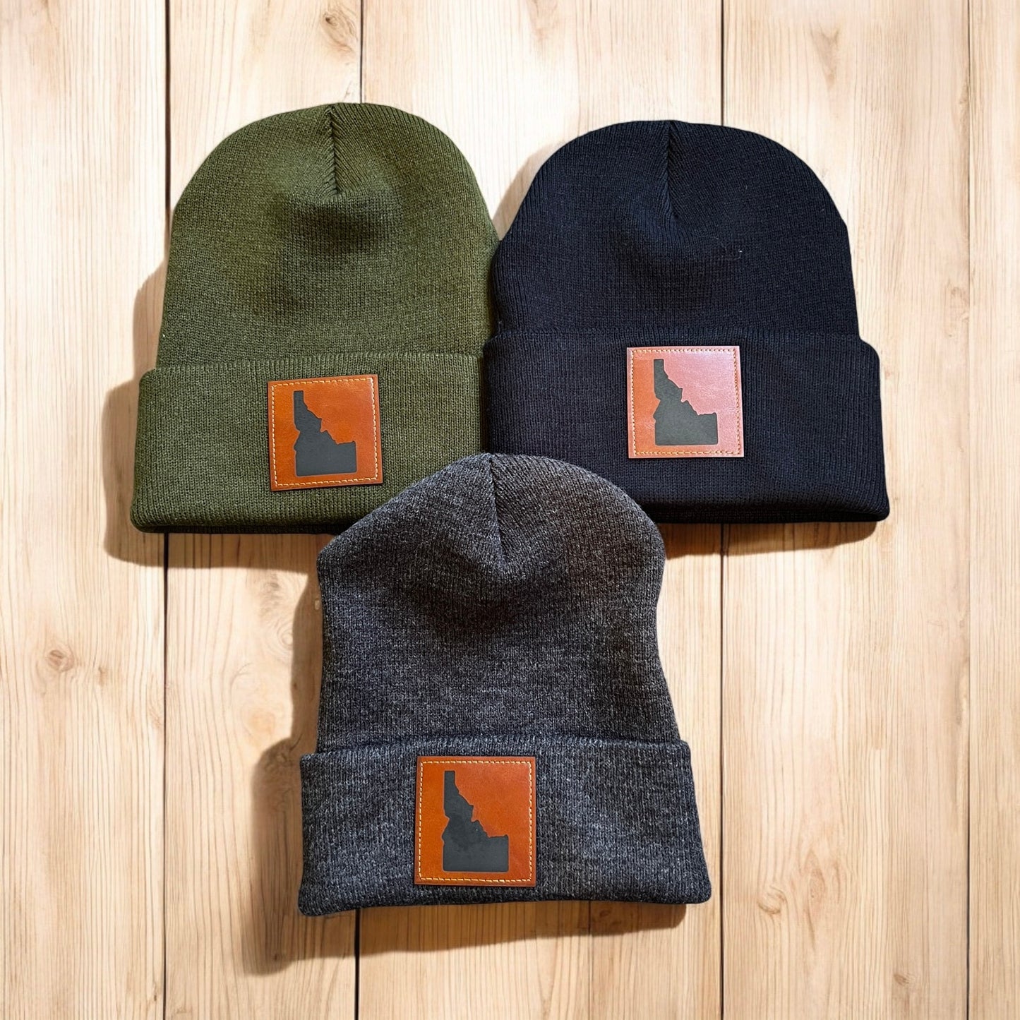 Three Idaho beanies are laying on a flat surface. The Idaho beanies are gray, black, and olive. Each beanie has a leather patch on it with Idaho. This Idaho hat will keep you cozy and warm all winter. This beanie is designed and made by TatorJo, an Idaho apparel company.