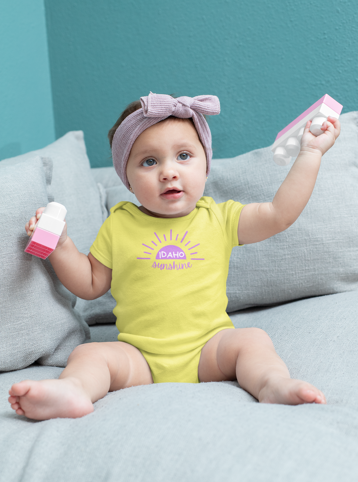 A little baby girl in an Idaho onesie that say Idaho Sunshine. The baby is wearing a bright yellow onesie. Printed by TatorJo.