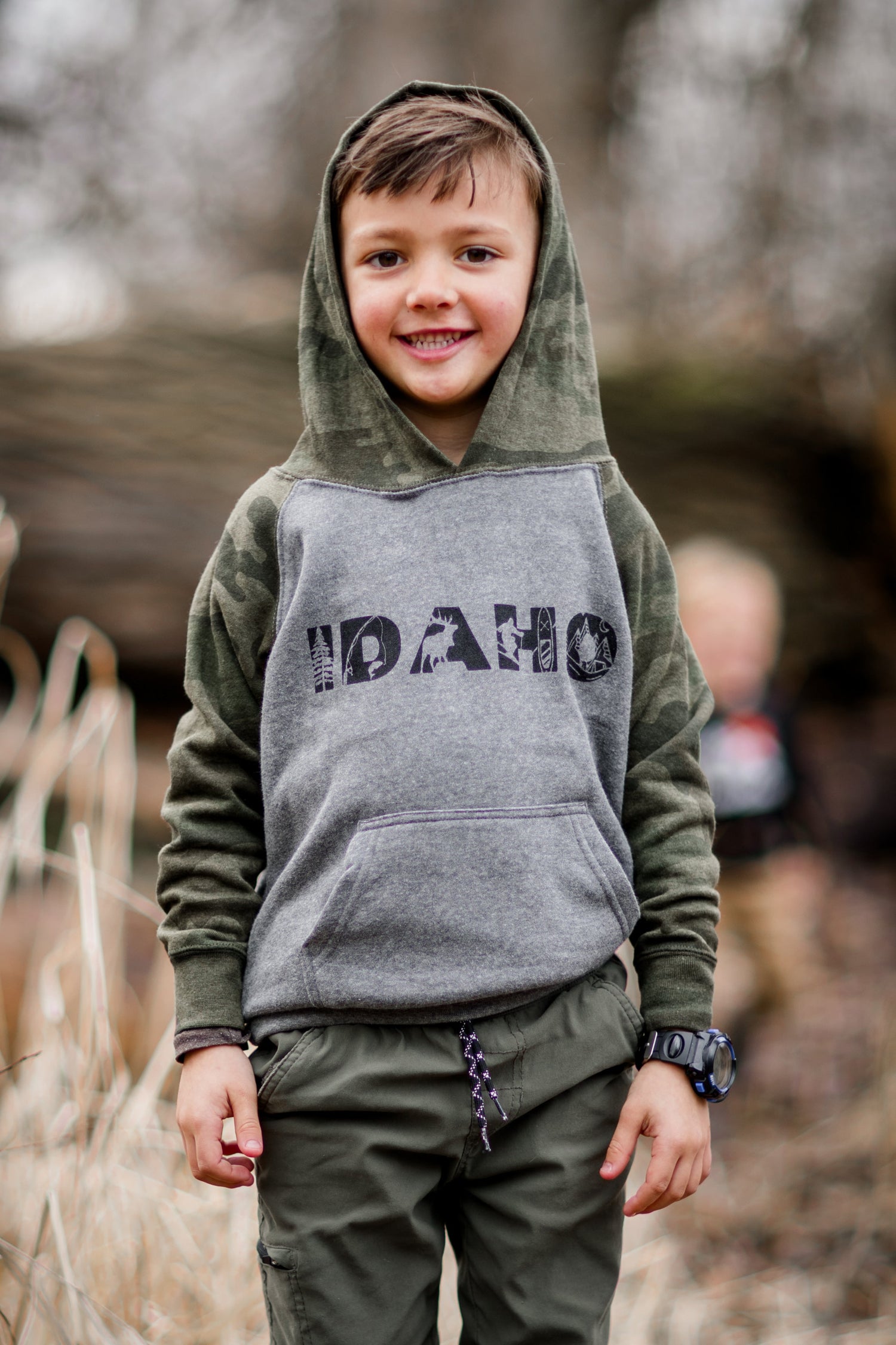 This Idaho boy is standing outside in Idaho wearing an Idaho sweater. The sweater had graphic designs on it that represent Idaho. The color is camo. 