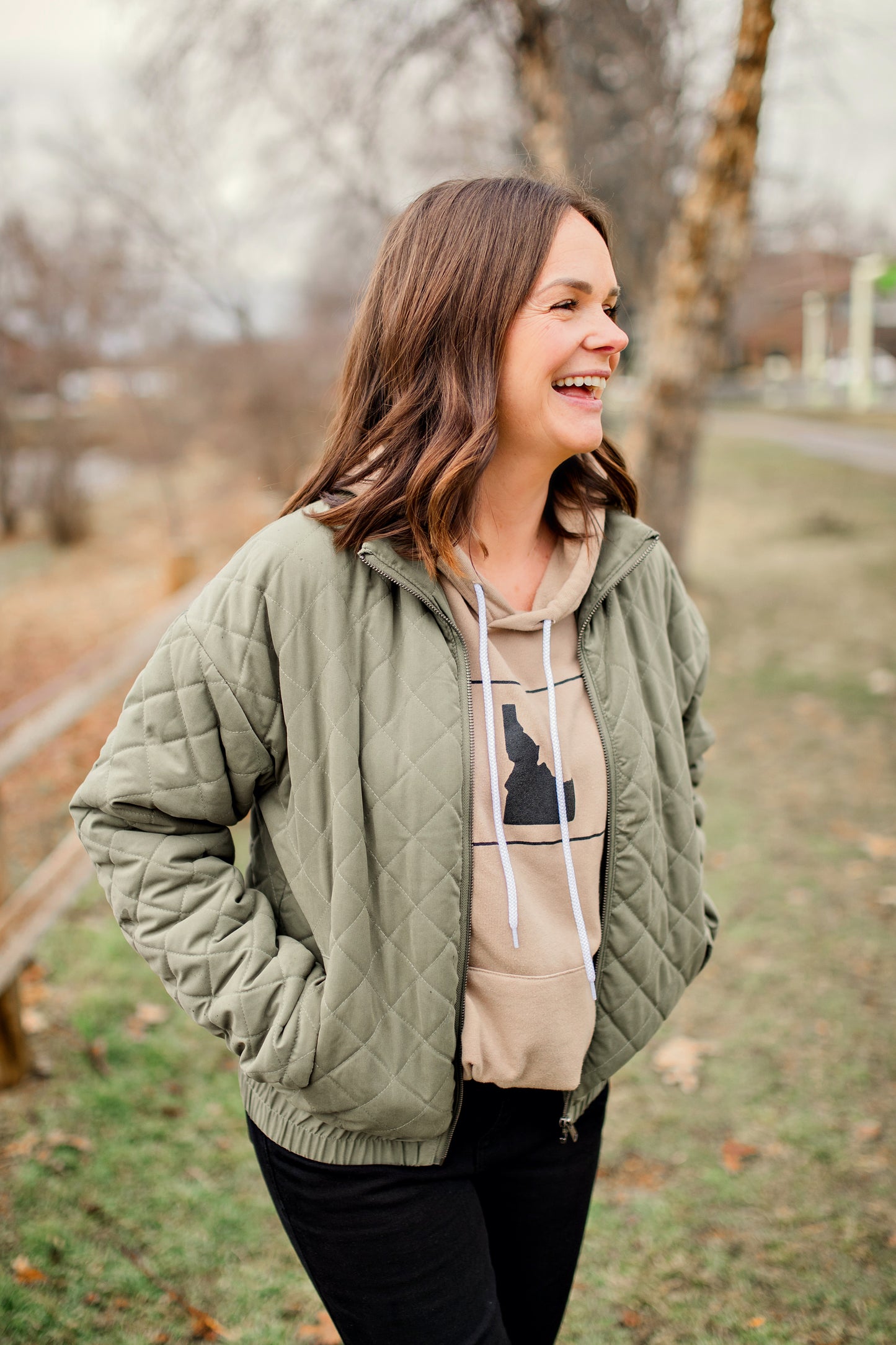 An Idaho mama wearing a tan Idaho hoodie. The hoodie is nicknamed the coziest hoodie in Idaho because it really is that coziest. This tan hoodie has a simple Idaho design on it. 