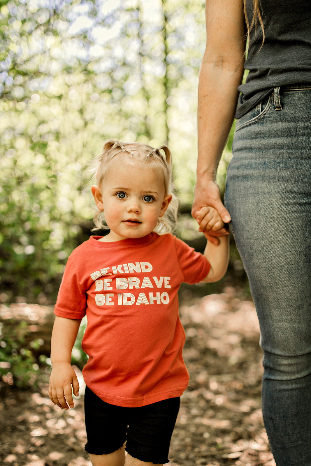 A little girl wearing a shirt that says Be Kind, Be BRave, Be Idaho on it. The little idaho girl is holding her moms hand. 