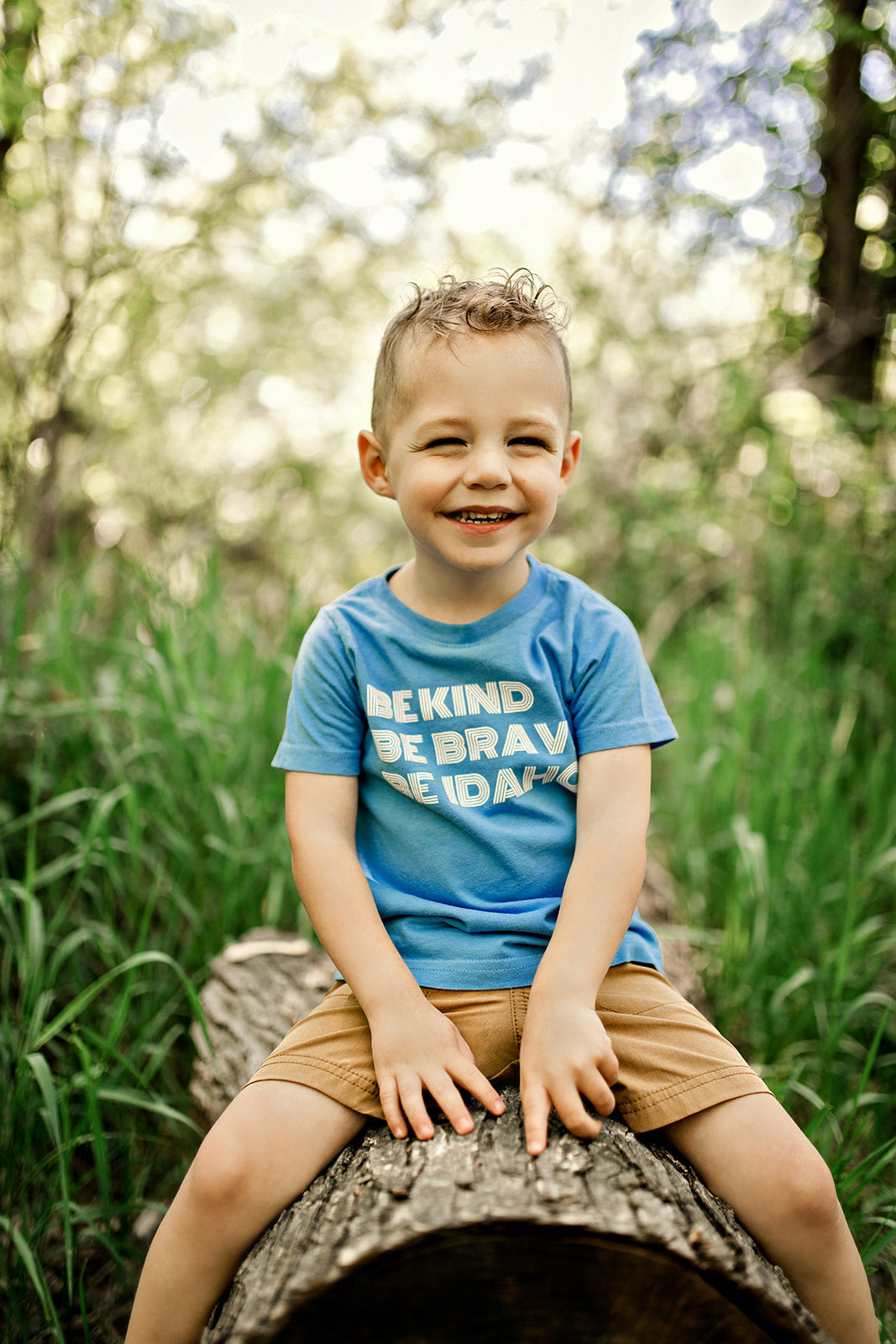 A little Idaho boy wearing an Idaho shirt that says Be Kind, Be Brave, Be Idaho on it. The shirt is in a bright blue color, the perfect Idaho Summer Tee.