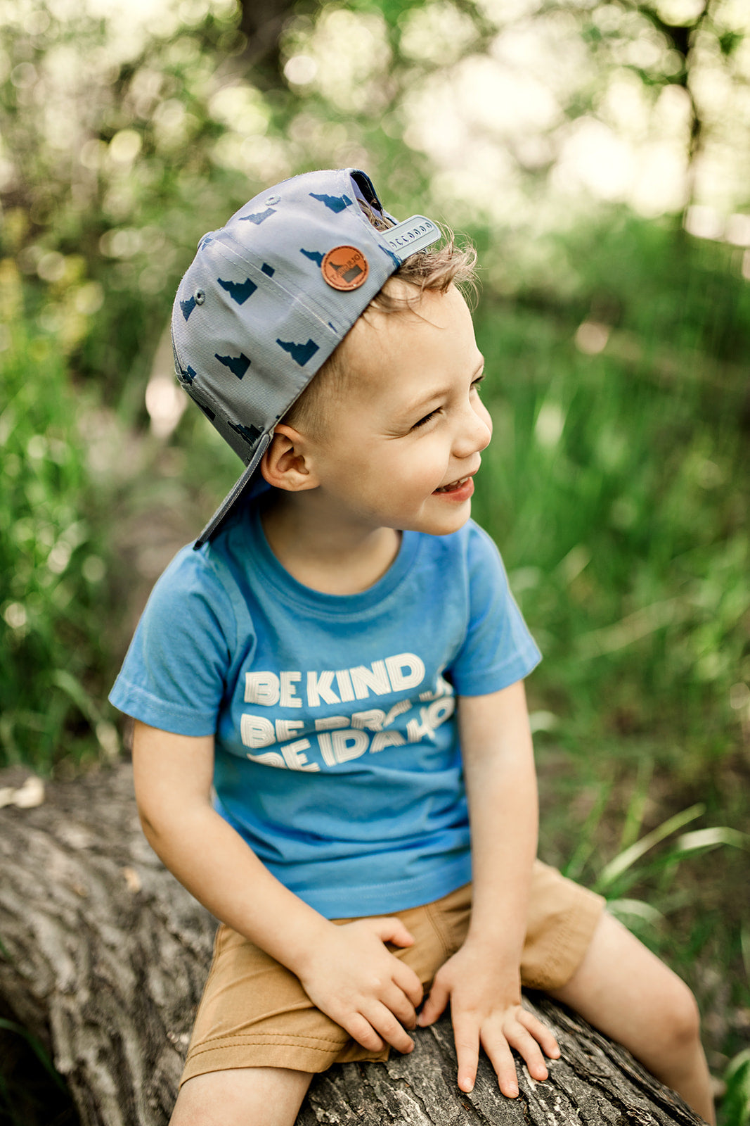 A young boy sitting on a log wearing a t shirt that says Be Kind, be Brave, Be Idaho on it. The T shirt is the perfect summer tee for those Idaho summers.