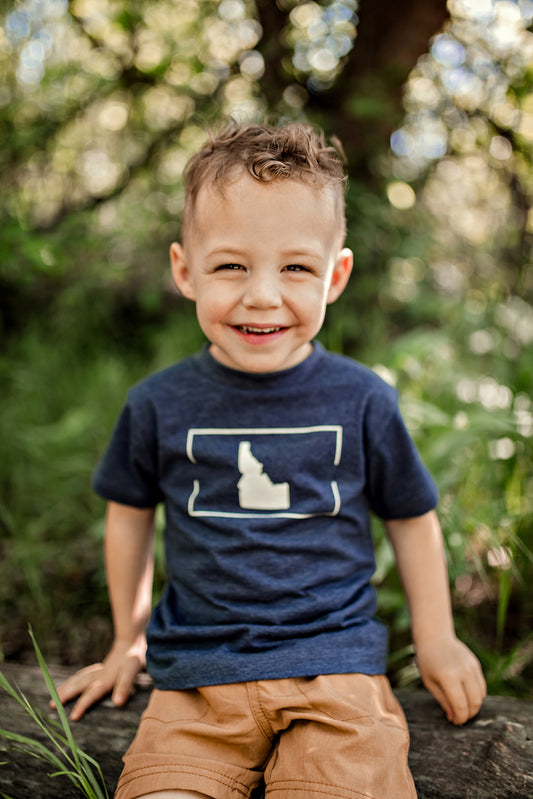 Cheerful young boy wearing an Idaho T-shirt and cap, smiling brightly in a sunlit forest. His joy highlights the adventurous spirit of Idaho's nature.