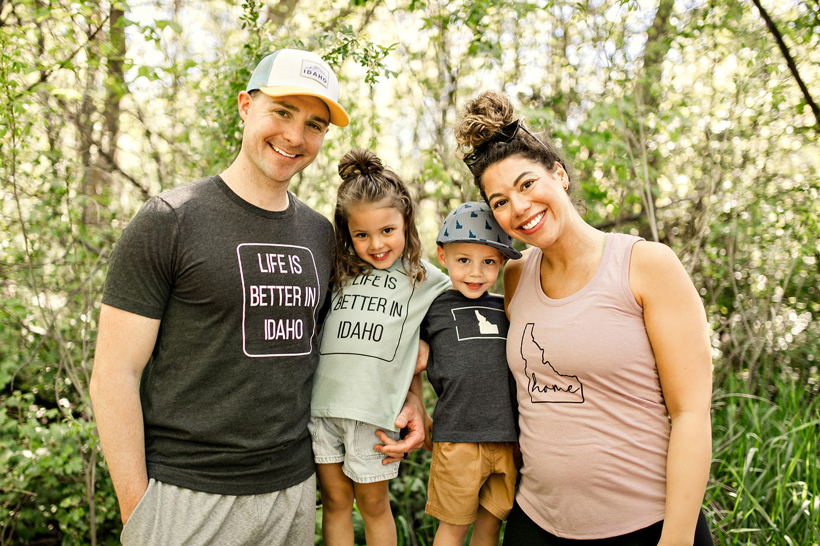 This picture showcases all the products TatorJo has to offer. It has the Mauve Idahome tank top and also the Life Is Better in Idaho, and other T shirts for kids. TatorJo is an Idaho Apparel company focused on providing apparel for Idaho Families. This picture is an Idaho family wearing TatorJo.