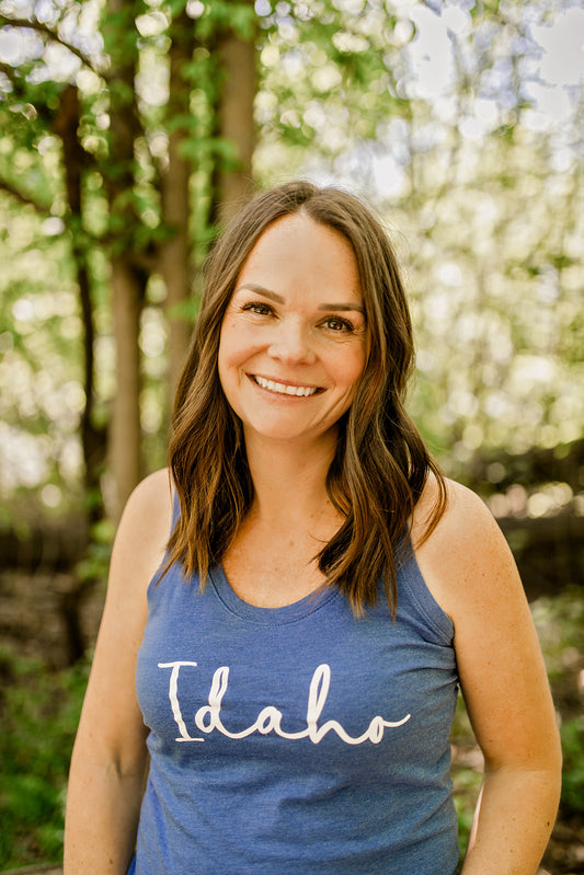 This racerback tank top has Idaho on the front, designed with a simple and elegant design that can be dressed up and dressed down. The blue tank top is made of triblend, which is ultra soft and durable. The classic design is screen printed in Idaho. This Idaho tank top is made by TatorJo, an Idaho Apparel Company. The women wearing the tank top is standing outside in Idaho.