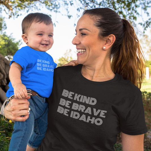 The mom is wearing an Idaho T Shir that says Be Kind, Be Brave, Be Idaho. This T shirt is black and the baby she is holding is wearing the same Idaho T Shirt. The Idaho Mama is standing outside. 