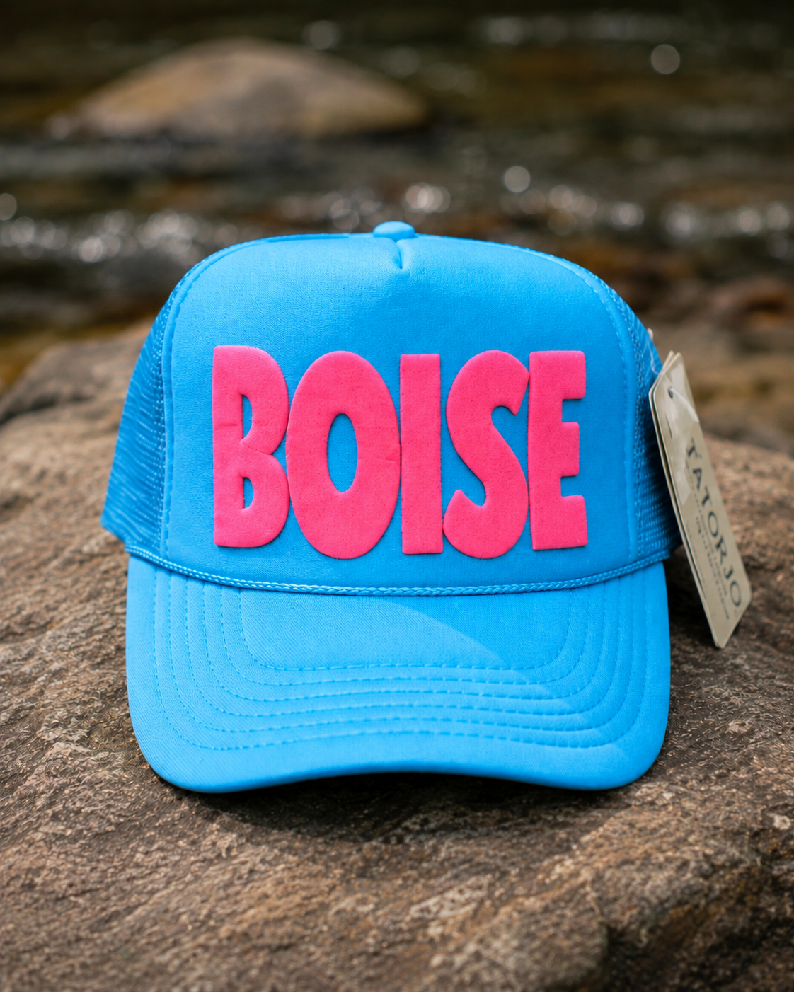 Blue cap with pink 'BOISE' text on a rock with water in the background