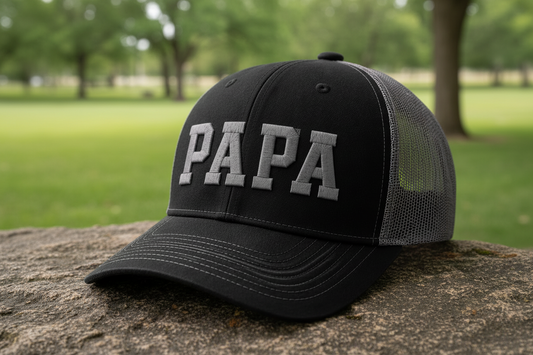 Black cap with 'PAPA' on a stone surface with a park background