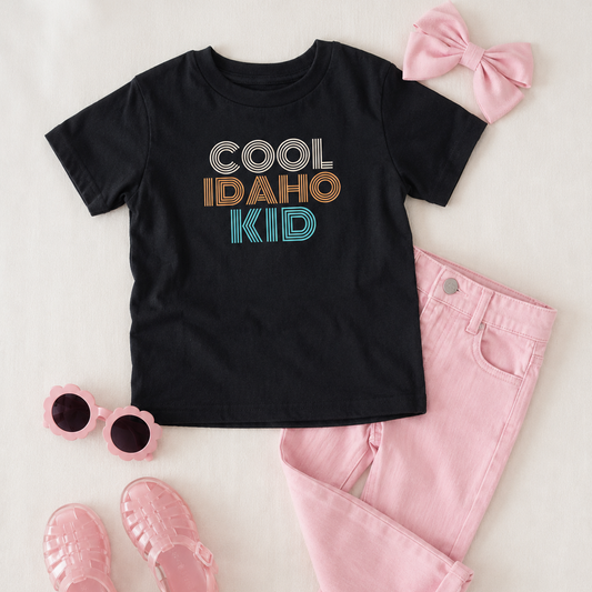 Black t-shirt with 'Cool Idaho Kid' text, pink pants, sunglasses, and sandals on a light background