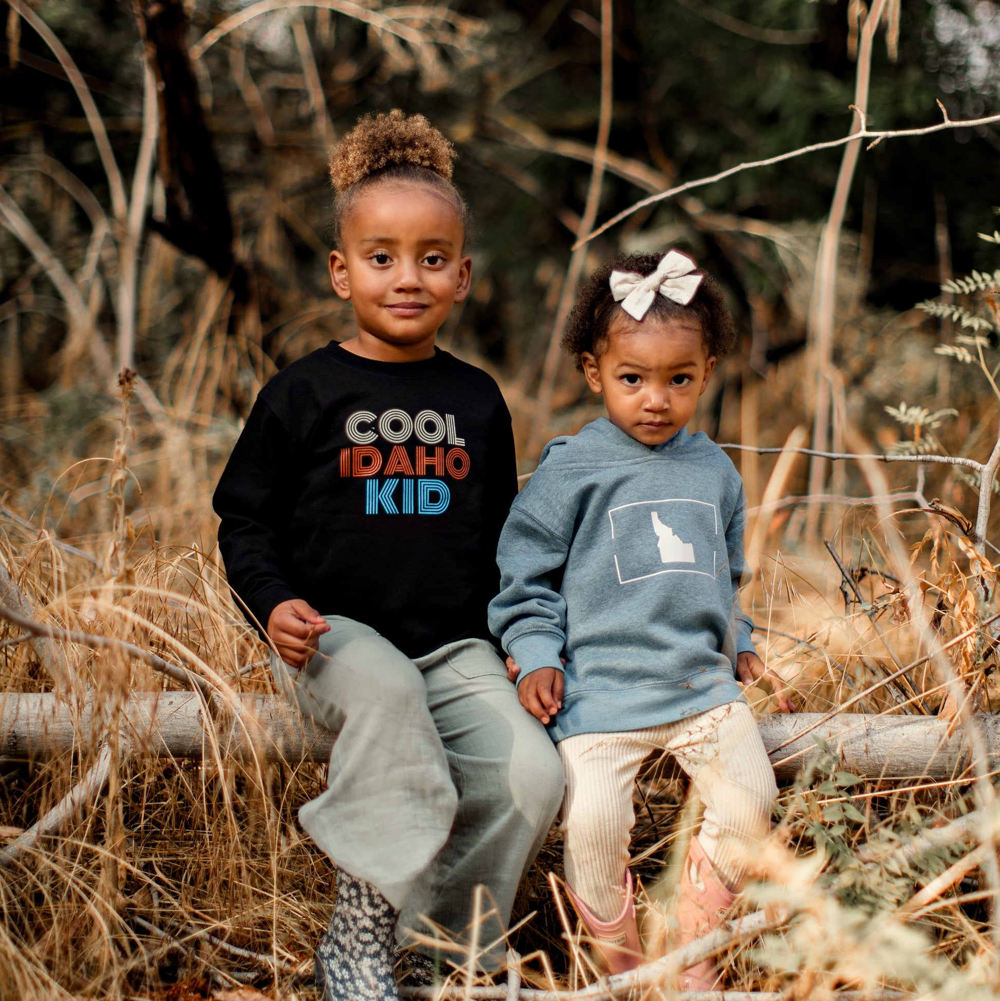 This adorable photo shows two little Idaho girls sitting outside. They are both wearing Idaho sweatshirts. One sweatshirt says Cool Idaho Kid on it. The other sweatshirt is a simple Idaho design. The sweaters are designed by TatorJo, an Idaho apparel company
