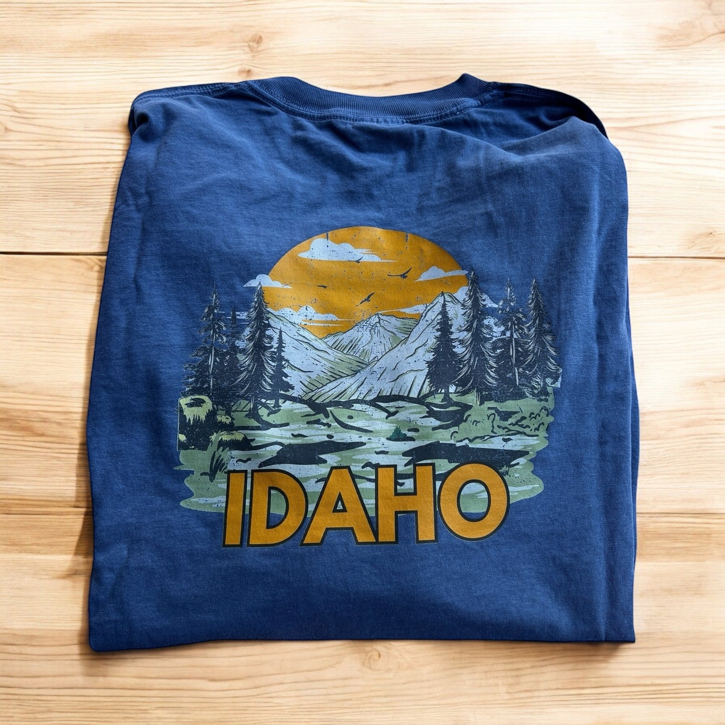 Blue Idaho Tshirt that has an image of mountains and a sun. The word has Idaho written on it. THere is a front corner and back image. This shirt is designed by TatorJo, an Idaho Clothing Company