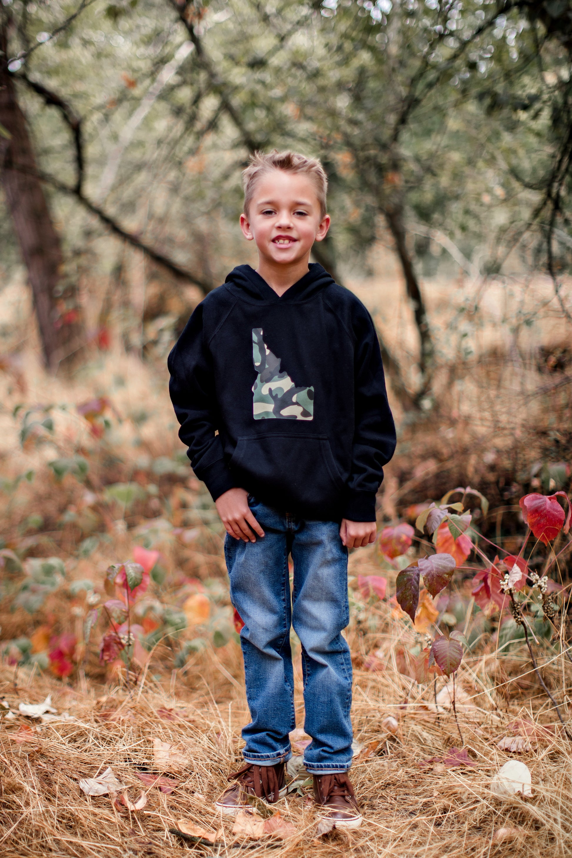 A young boy standing outside in Idaho wearing a black hoodie with Idaho on it. The Idaho has a camo inside of it. The design is created by TatorJo, an Idaho based apparel company.