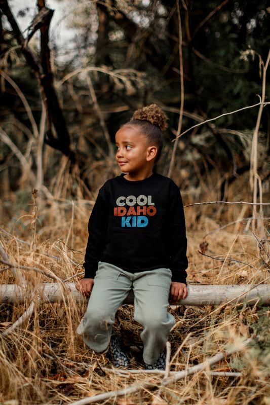 A little girl sitting outside in Idaho wearing Idaho clothing. The Idaho sweater says Cool Idaho Kid. This sweater is designed by TatorJo, an Idaho apparel company