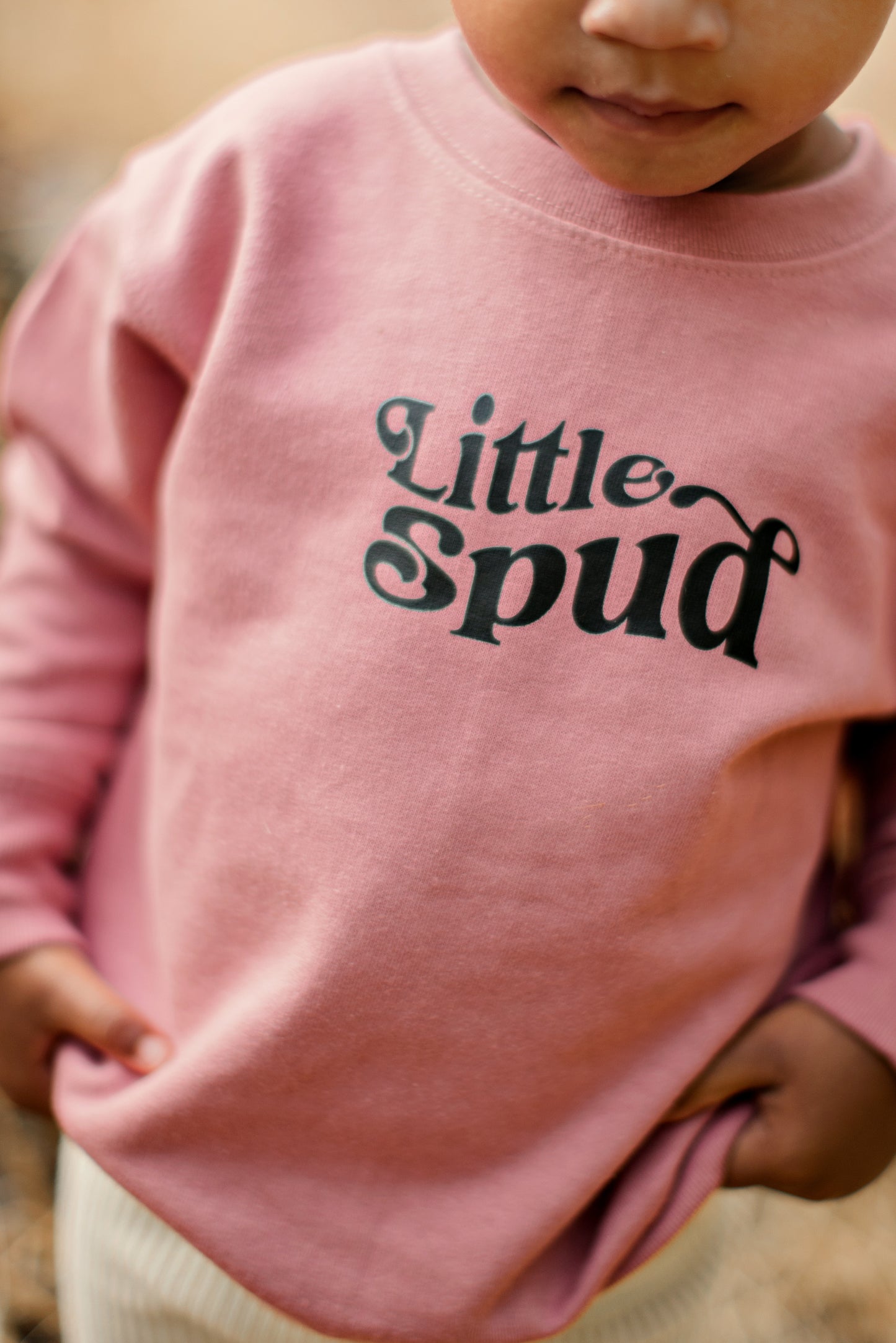 Mauve pink sweater that says Little Spud, The Little Spud design represents Idaho and the spuds we offer. The little girl is outside in Idaho