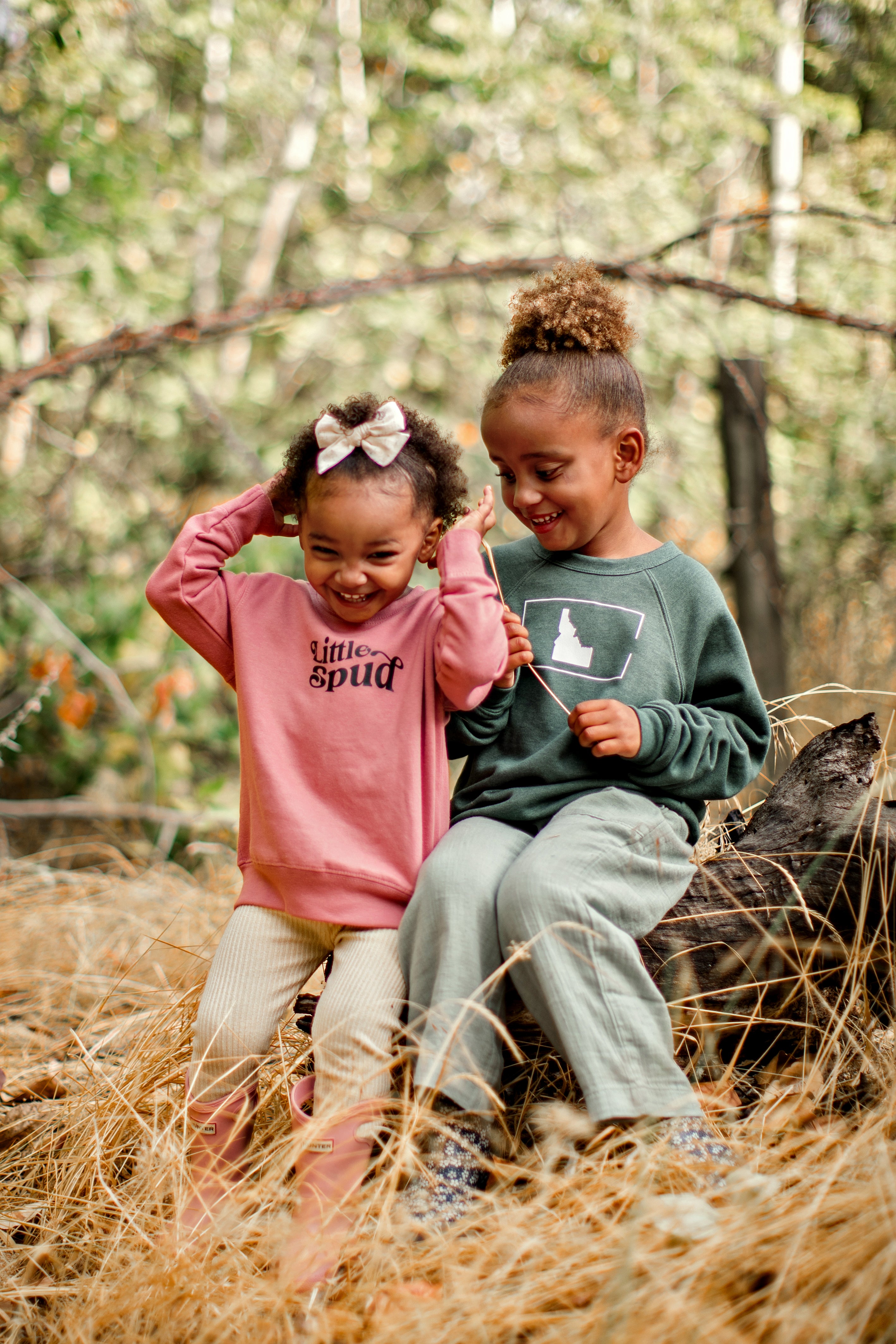 Two little Idaho girls hanging outside in their adorable TatorJo sweaters. One sweater is green with an Idaho on it and the other is a mauve Little Spud sweater. Both are designed and printed in Idaho by TatorJo