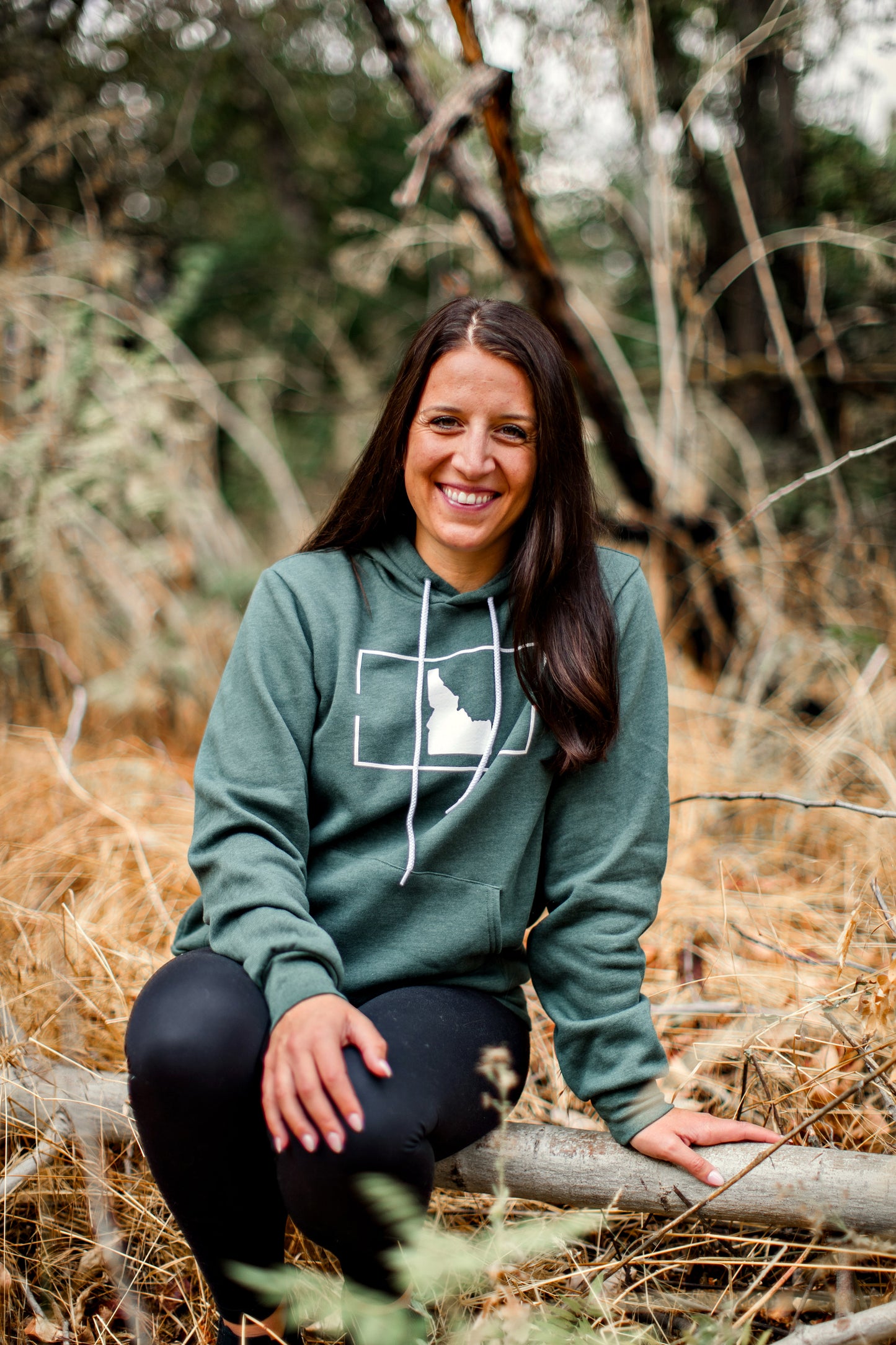 A young mom wearing a forest green hoodie that has a simple Idaho design on it. The Idaho sweatshirt is so cozy and soft. This sweatshirt comes with a hood, like a hoodie. It is designed by TatorJo, an Idaho Clothing Business