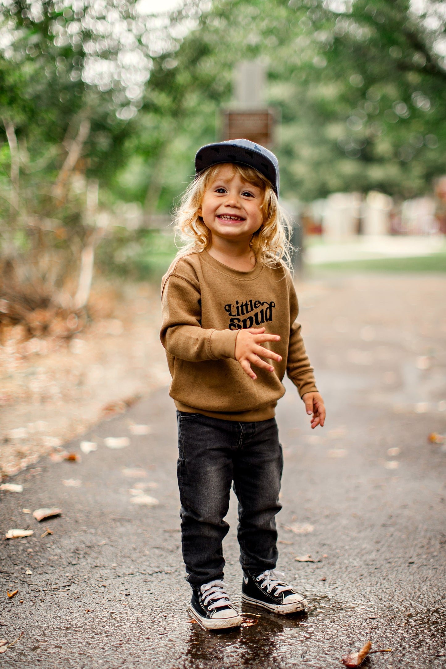 an Idaho sweater that says Little Spud on it, the sweater is a brown. The little boy is wearing an Idaho hat that has little blue Idaho's all over it. The sweater is an Idaho sweater that is designed by TatorJo, an Idaho apparel company