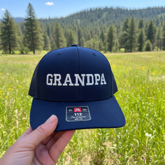 Navy blue cap with 'GRANDPA' text held by a hand against a wooden background
