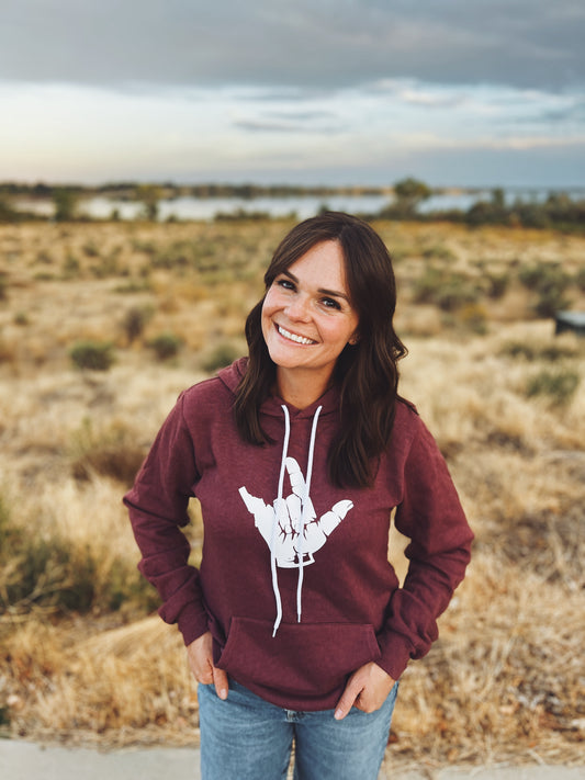 Woman wearing a maroon hoodie with a white graphic in an open field. This Idaho Hoodie has Idaho printed on the hoodie inside the sign language sign