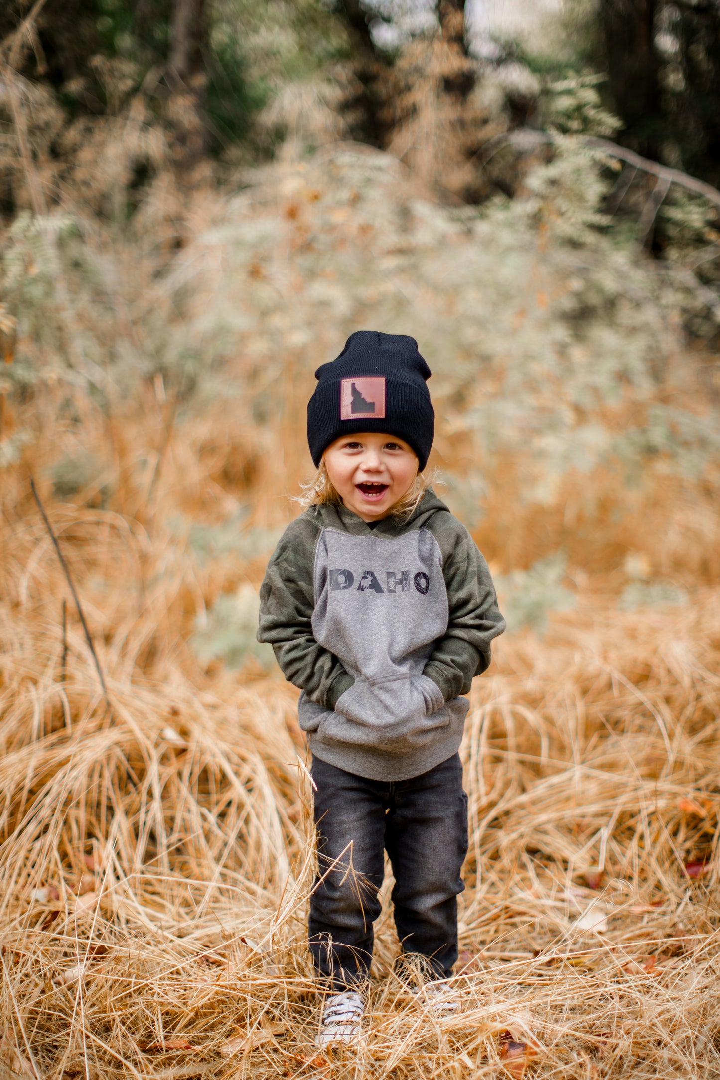Little Idaho boy wearing an Idaho sweater. The sweater has the word Idaho on it with graphics that represent Idaho. The sweater is a camo color. He is wearing an Idaho beanie, black Idaho beanie with a leather patch. The patch has Idaho on it. 