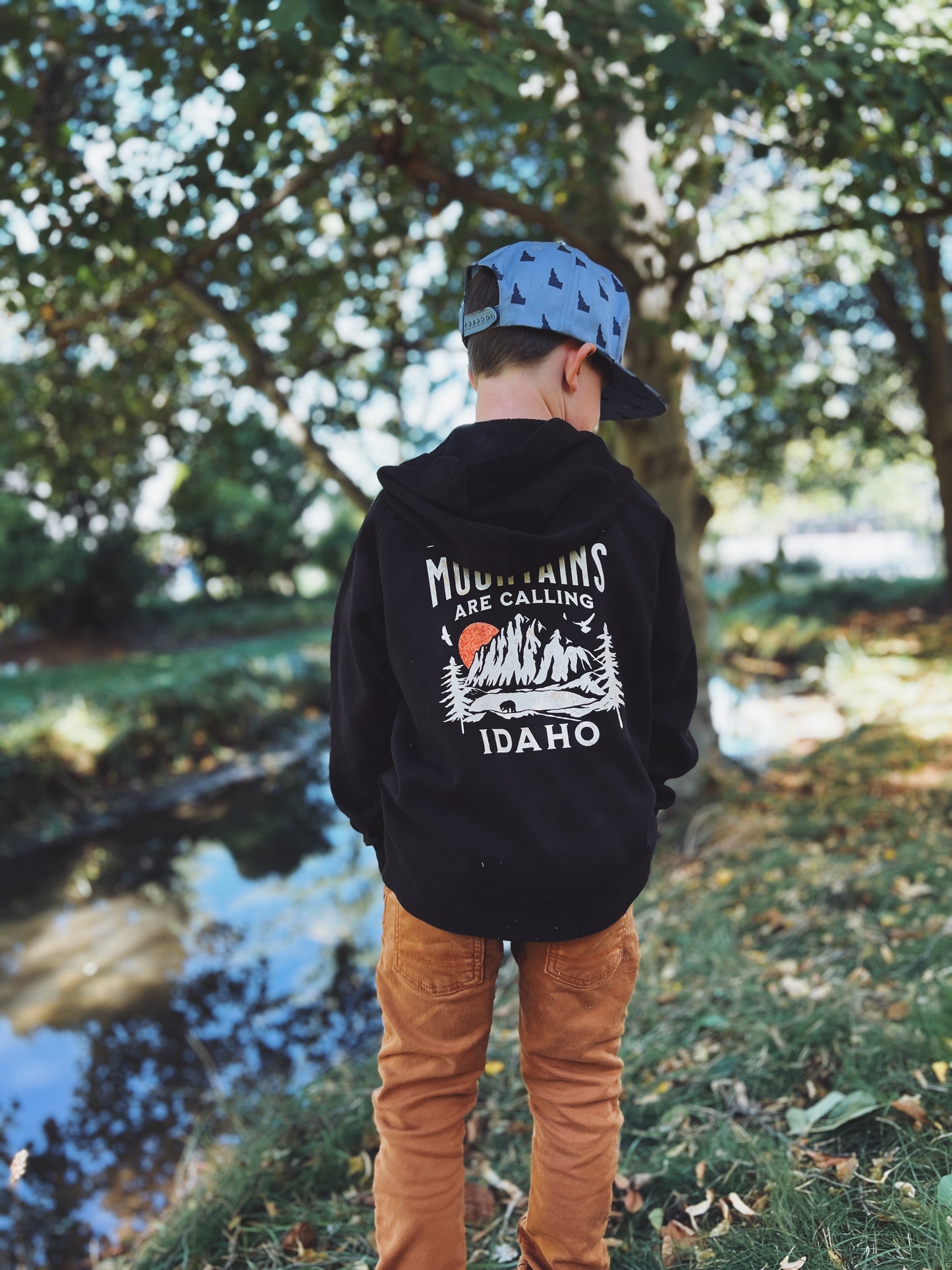 Child wearing a black Idaho hoodie with text and graphics, standing outdoors near a body of water. The Hoodie reads, Mountains are Calling Idaho, this hoodie is a perfect adventure hoodie while living in Idaho